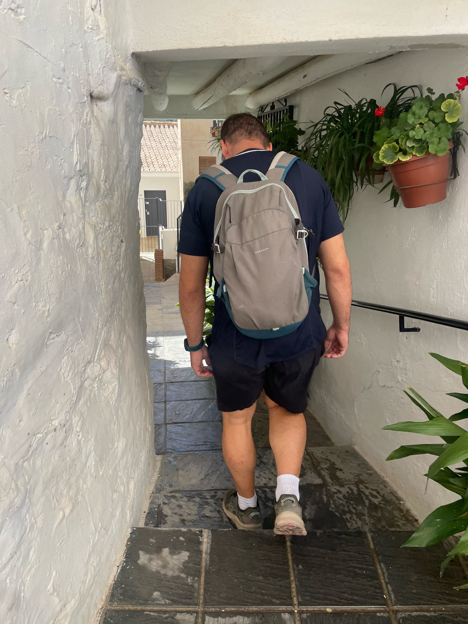 A man walks down a narrow, sloped alleyway in Güéjar Sierra, Spain. He is seen from behind, wearing a dark blue t-shirt, black shorts, white socks, and hiking shoes. A light gray backpack with blue straps is on his back. The alley walls are rough-textured and painted white. A black handrail is attached to the right wall. Potted plants with green leaves and red flowers hang on the right wall. The ground is paved with dark gray tiles. In the background, a building with red tiled roofs is visible.