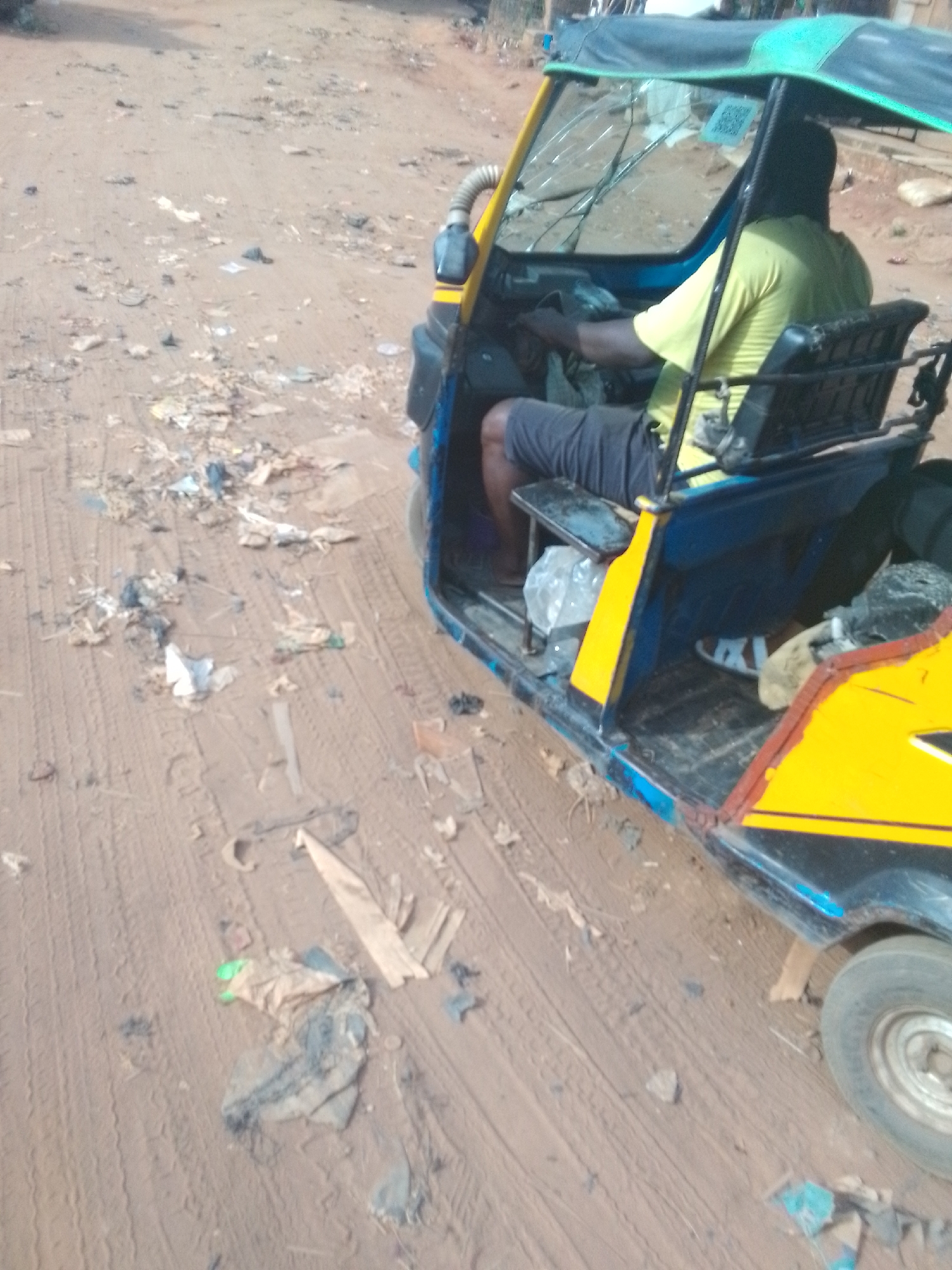 The image shows a three-wheeled motor vehicle, commonly known as a "tuk-tuk" or "auto-rickshaw", parked on a dirt road. The tuk-tuk is painted in blue and yellow and has a partially broken windshield. A person wearing a yellow shirt and dark shorts is seated inside, presumably the driver. The road is littered with trash. The location appears to be an unpaved street in the city of Unknown, Unknown.
