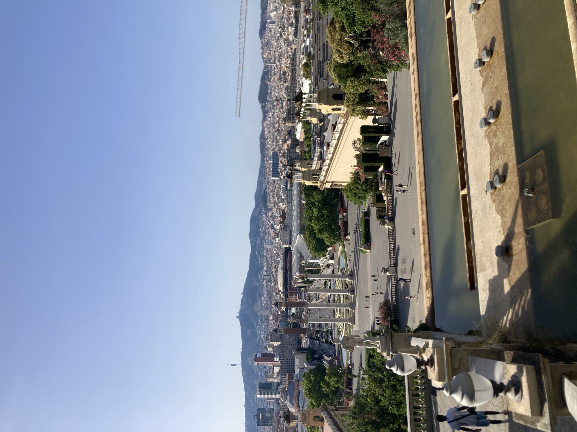 This is an outdoor, elevated shot of the Montjuïc hill in Barcelona, Spain, looking out over the city. In the foreground, there are two decorative lamps on a stone balustrade, and beyond that, a partially visible shallow fountain with some spray nozzles. In the middle ground, there is a wide plaza with several people walking, and a row of large columns. In the background, the city of Barcelona stretches out, with a prominent communications tower on a hill in the distance. A large construction crane is visible in the far right background. The sky is clear and blue.