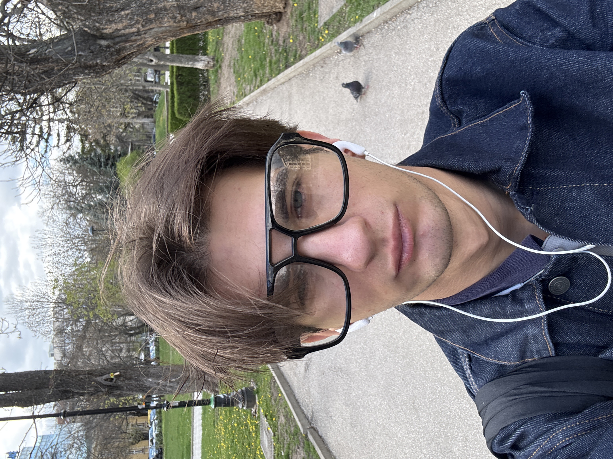 A young man is taking a selfie on a path in a park. He is wearing a dark blue denim jacket over a dark shirt, eyeglasses with thick black frames, and white earbuds. The park has trees, grass, and a paved walkway where two pigeons are visible. Based on the architectural style and vegetation, the location is likely in Sofia, Bulgaria.