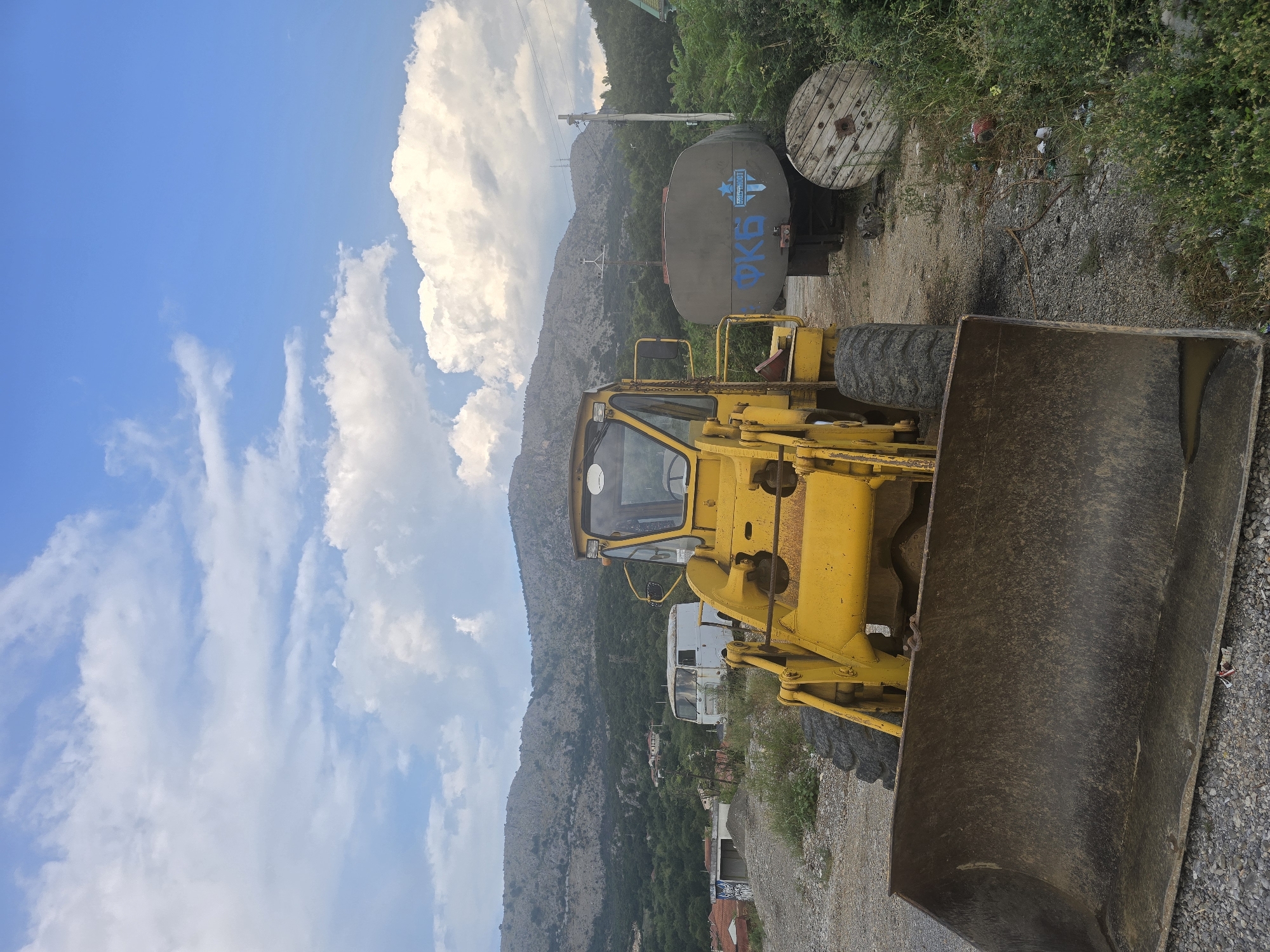 The image depicts a yellow front loader parked on a gravel lot, facing the viewer. The loader's bucket is large and appears worn. Behind the loader, a grey tanker trailer with blue graffiti on its side is visible, along with a large wooden cable spool. In the background, a white van and some houses are scattered amongst dense green foliage, with a rugged, treeless mountain range dominating the horizon. The sky is partly cloudy with white cumulus clouds against a bright blue backdrop. The scene appears to be an outdoor storage or work area, possibly on the outskirts of a settlement in Podgorica, Montenegro, given the mountainous terrain and vegetation characteristic of the region. There are no people visible in the image.