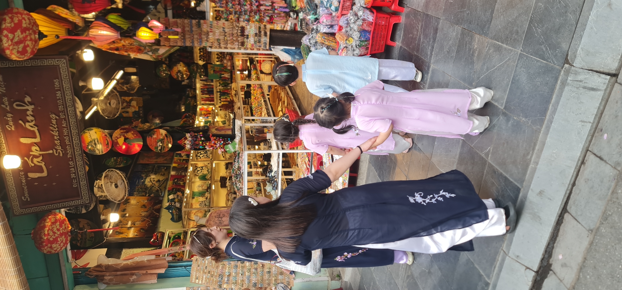 Here's a description of the image:
A group of people, including three children and two adults, are standing in front of a shop in Hội An Ward, Vietnam. The children are wearing traditional Vietnamese Áo dài dresses in light pink and light blue colors. The women are also dressed in Áo dài dresses. The children hold hands. The shop has various items for sale, including souvenirs, lanterns, and handicrafts, displayed on shelves and hanging from the ceiling. The shop's sign reads "Quay Lau Lấp Lánh Sparkling Souvernier, 39 Lê Lợi - Thành phố Hội An- ĐT: 0510.386 1065". The street is paved with stone tiles.
