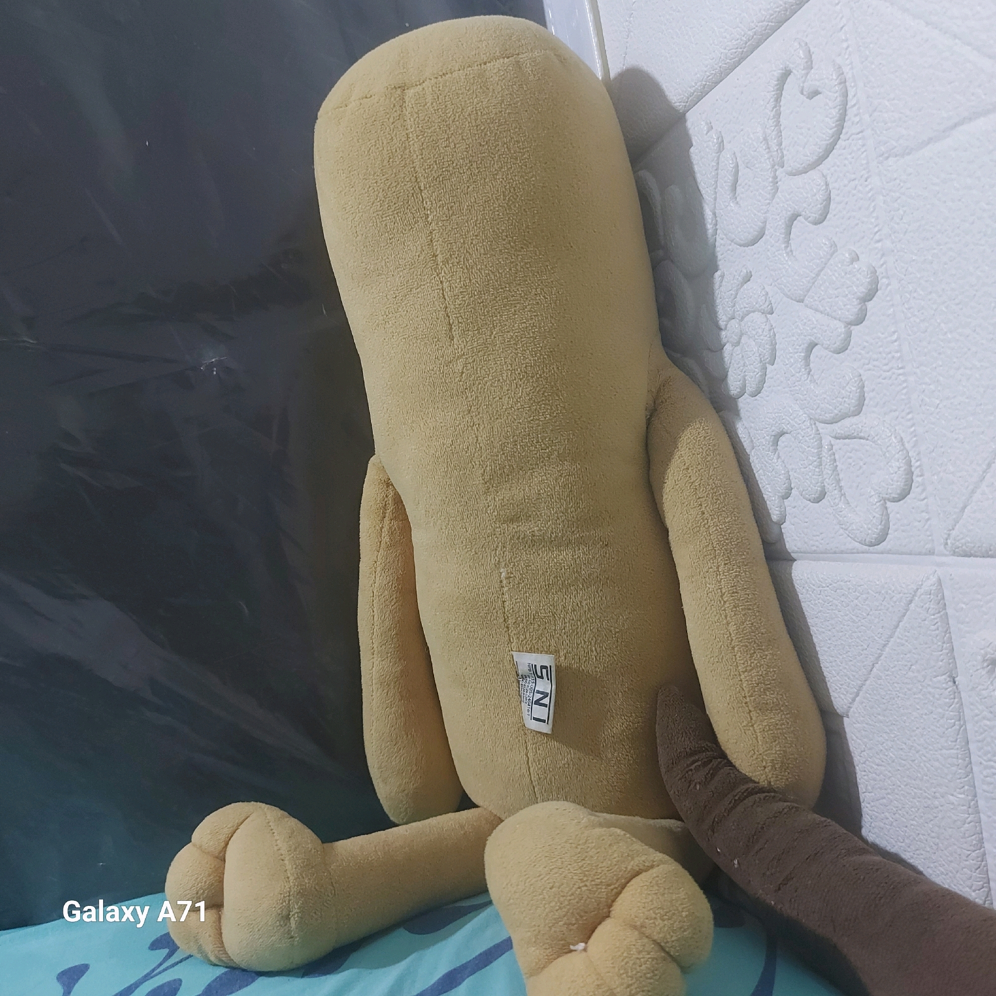 A plush toy, predominantly yellow, is positioned on a blue and white striped surface. The toy is seen from the back, leaning against a textured white wall with an embossed floral pattern. A brown tail-like appendage extends from the lower right side of the toy. A white tag with the letters "SNI" is affixed to the back of the toy. The background on the left consists of a dark, somewhat shiny surface that appears to be plastic sheeting or a similar material. The lower left corner of the image displays the text "Galaxy A71". This scene is likely a domestic setting in Surabaya, Indonesia.