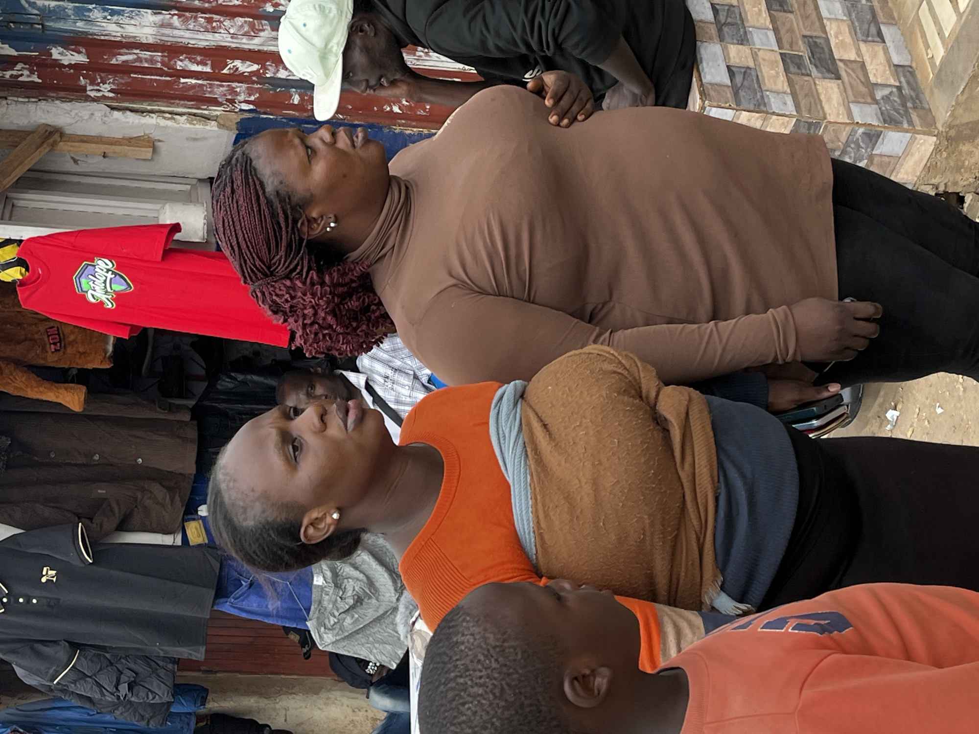 In Gura Top, Nigeria, two women and a child stand outside a shop displaying clothing. The woman on the left wears an orange shirt and a brown wrap. The woman on the right is in a brown turtleneck and dark pants. A young child in an orange shirt stands to the left. Behind them, a variety of clothing items hang, including a black polo shirt and a red t-shirt. A man in a light green cap and a black hoodie is visible in the background. The shop appears to have a corrugated metal facade and a tiled counter.