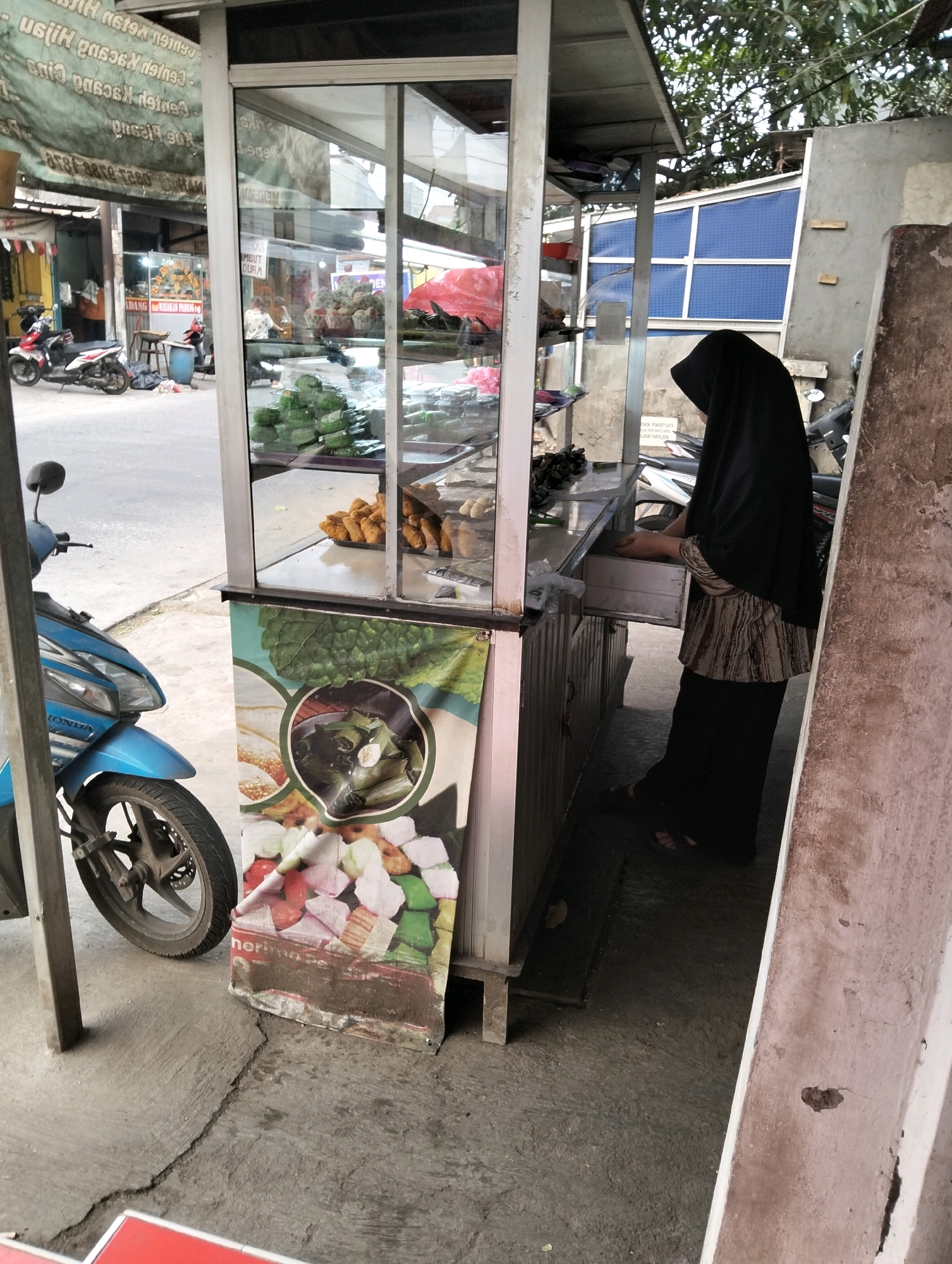 In the city of Cileungsi, Indonesia, a street food vendor is captured at work in front of a small, glass-enclosed display cart. The vendor, dressed in a black hijab and a patterned long-sleeved shirt, is seen interacting with the cart's drawers. The cart itself is filled with various food items, including what appears to be fried snacks and green, wrapped snacks. A blue motorcycle is parked to the left of the cart. In the background, a banner with images of food items is displayed on the front of the cart. The scene is set on a concrete ground, with a building and trees visible in the distance.