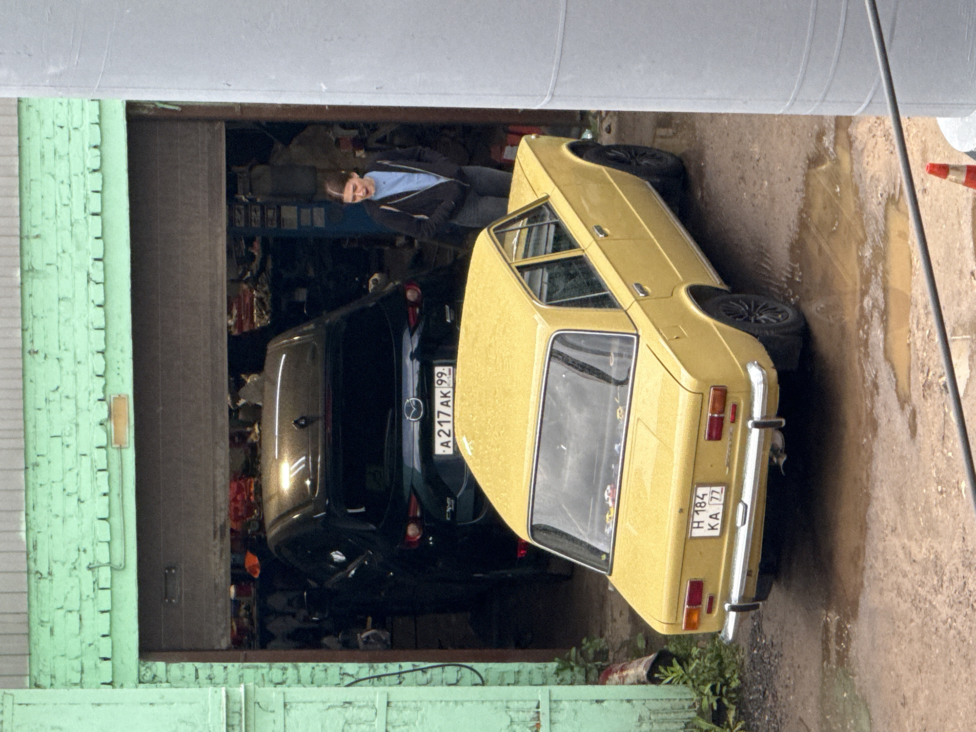 In Moscow, Russia, a young woman with brown hair tied back stands in the doorway of a garage, wearing a black zip-up hoodie over a light blue t-shirt and dark jeans. Her mouth is open, as if speaking or exclaiming. In front of the garage, a vintage yellow sedan, appearing to be a VAZ-2101, is parked on a muddy, wet surface. The car has chrome bumpers and trim, and a license plate reading "H 184 KA 77". Inside the garage, partially visible, is a dark-colored SUV, likely a Mazda. The garage entrance is framed by light green brick walls. The scene appears to be outdoors, possibly in a service area or a private yard, with puddles of water suggesting recent rain.