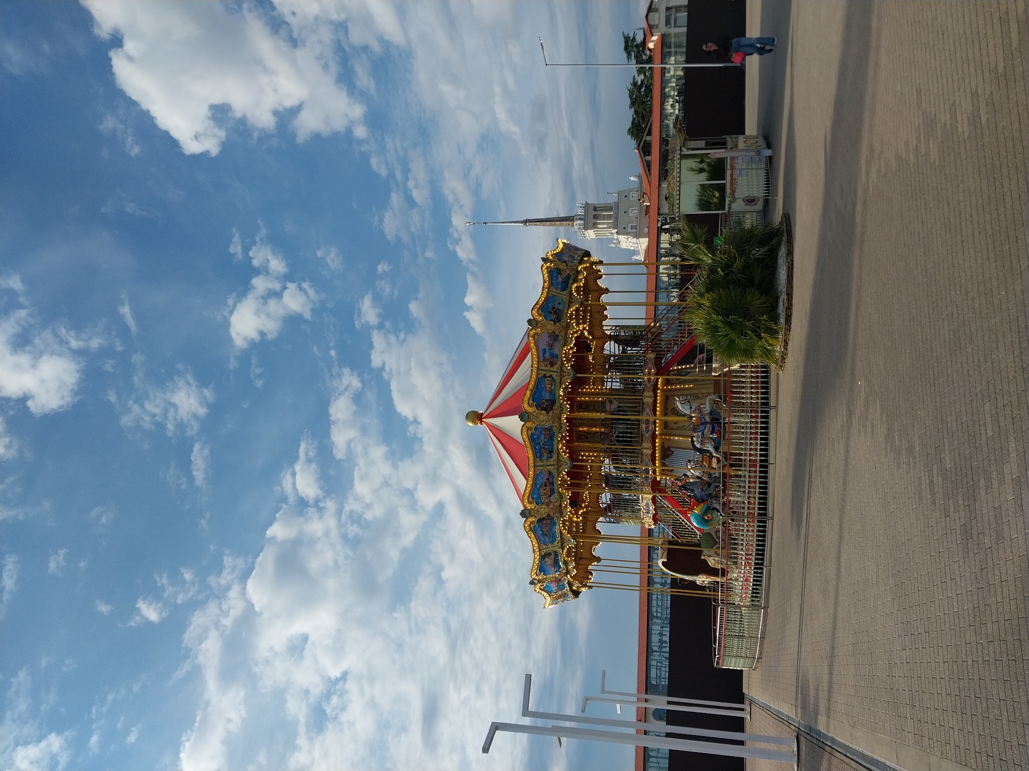 The image captures an outdoor public space in Sochi, Russia, dominated by a brightly decorated carousel under a clear blue sky.
The carousel, positioned towards the center-left, is ornate with golden accents, colorful painted panels depicting scenes, and a striking red and white striped canopy. Numerous small lights adorn its structure, and it is surrounded by a metal safety fence.
In the background, a prominent white building with a tall, slender spire stands against the sky, characteristic of the Sochi Marine Terminal. Other buildings with red-tiled roofs and reflective windows are also visible, suggesting a waterfront or urban resort setting.
The foreground and right side of the image feature a wide, clean paved pedestrian area made of light-colored interlocking bricks, extending into the distance. A small, bushy, palm-like plant grows in a planter box near the carousel. Further back on the paved path, a single person, dressed in dark clothing and blue jeans and carrying a red bag, walks away from the viewer.
The sky occupies a significant portion of the upper frame, displaying a vibrant blue with scattered, fluffy white cumulus clouds, indicating bright and fair weather. Streetlights and white structural posts are also present in the scene. The overall impression is one of a lively and well-maintained public area on a pleasant day. No discernible text is visible.