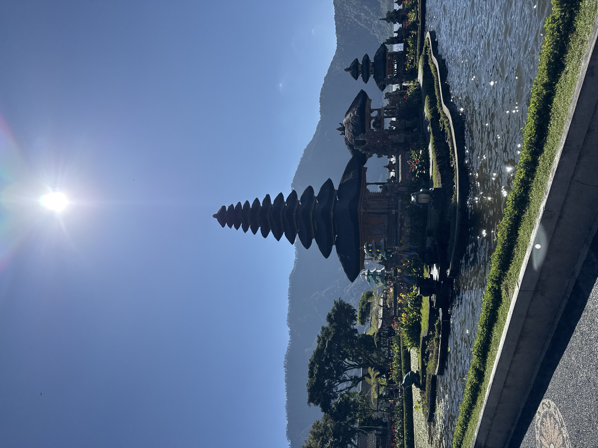 The image depicts the Pura Ulun Danu Bratan temple complex, a serene water temple located in Baturiti, Bali, Indonesia. The temple is situated on a small island in the middle of Lake Bratan, surrounded by lush greenery and a backdrop of majestic mountains. The sun shines brightly in the clear blue sky, casting a warm glow on the scene and creating sparkling reflections on the water.
The main structure of the temple is a towering meru with multiple tiers, a characteristic of Balinese Hindu architecture. Adjacent to it are smaller shrines and pavilions, each with its own intricate details and thatched roofs. The temple grounds are meticulously maintained, with vibrant flowers, manicured lawns, and ornate statues that add to the mystical ambiance.
Several small figures can be seen scattered around the temple complex, suggesting the presence of visitors or devotees. The calm waters of Lake Bratan surround the temple, providing a sense of tranquility and reflecting the beauty of the surrounding landscape. The image captures a moment of peaceful contemplation, inviting the viewer to immerse themselves in the spiritual and natural splendor of this iconic Balinese landmark.
