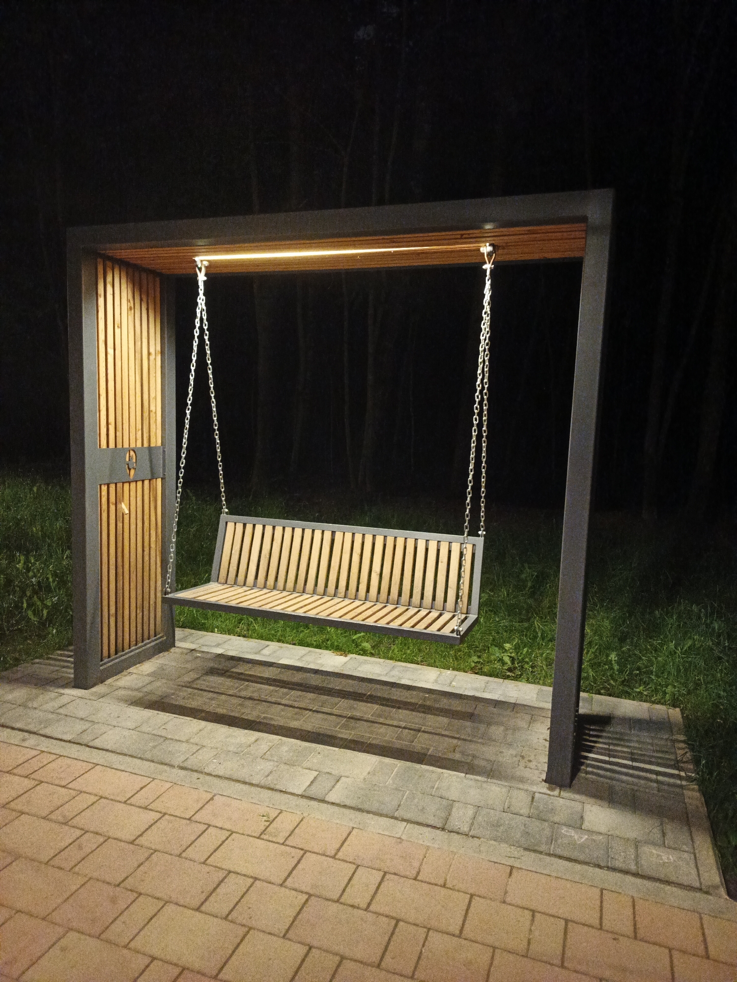 In Sof'ino, Russia, a modern, illuminated park bench swing is the central subject of this nighttime photograph. The swing, constructed from metal with a wooden slatted seat and backrest, hangs from a dark grey metal frame. Integrated warm LED lighting illuminates the top of the frame. The frame is supported by two vertical posts; one is adorned with vertical wooden slats and a small, circular emblem. The swing is situated on a paved area composed of interlocking brick pavers in shades of grey and orange, bordering a grassy area. In the background, a dark, out-of-focus forest serves as the environmental backdrop. The lighting casts distinct shadows of the swing's chains and frame onto the paved surface.