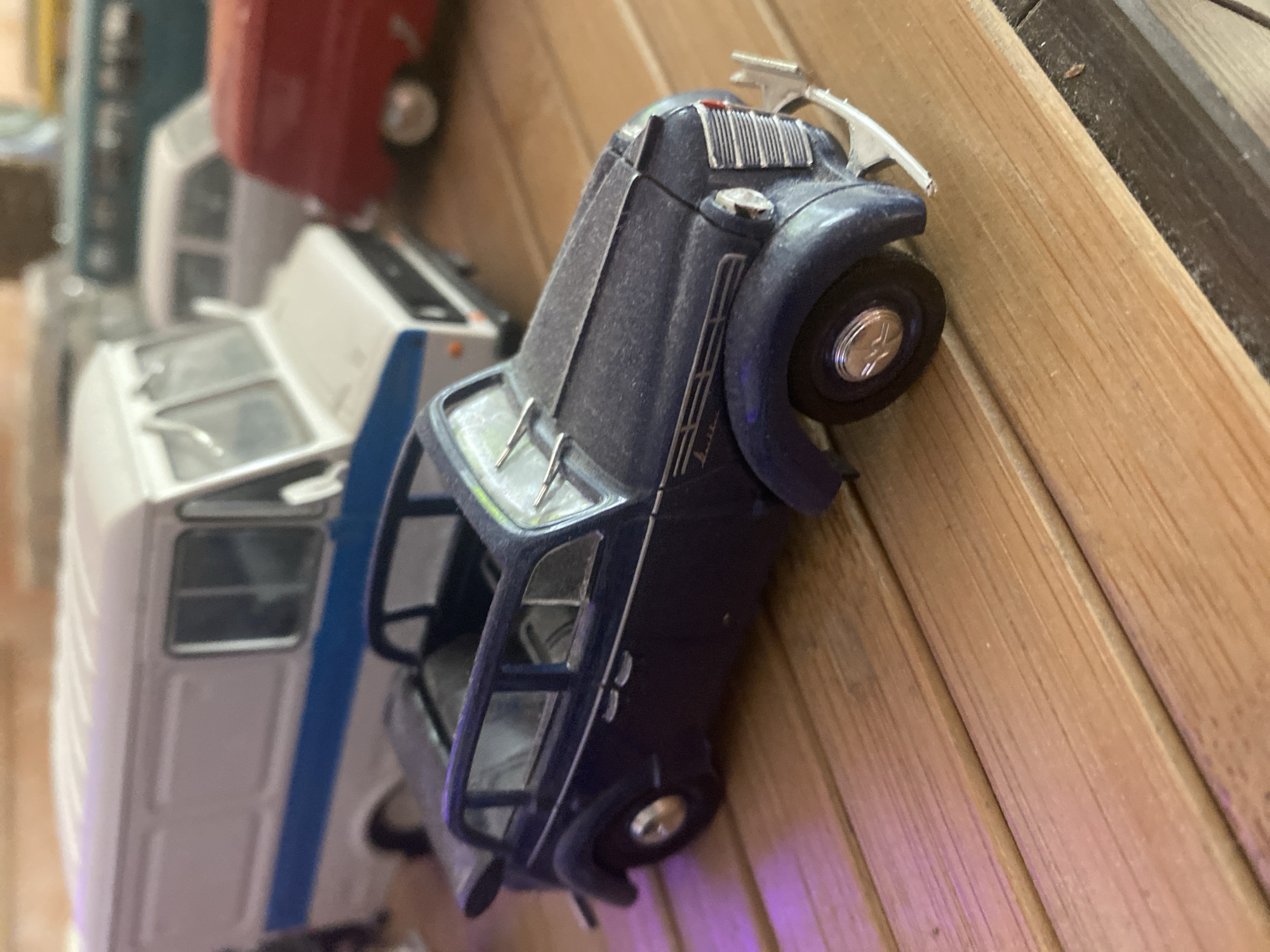 In a close-up, low-angle shot, a collection of die-cast model vehicles is displayed on a wooden surface. The primary subject is a dark blue sedan, positioned in the foreground with its front-left quarter visible. The model features detailed chrome accents, including the grille, headlights, door handles, and trim. The windows are clear, and wipers are present on the windshield. Behind the blue sedan, a white and blue van is partially visible. To the right of the van, a red van-like vehicle is also in view, with its front end obscured. Further in the background, other model vehicles, including a teal-colored bus, are faintly discernible. The wooden surface appears to be made of planks and has a warm, light brown tone, with some planks exhibiting a slightly darker hue. Pinkish light reflects from the left side of the blue sedan, suggesting a lighting source in that area. The overall setting appears to be indoors, on a shelf or display surface, rather than outdoors in a city environment. There are no people or animals present in the image.