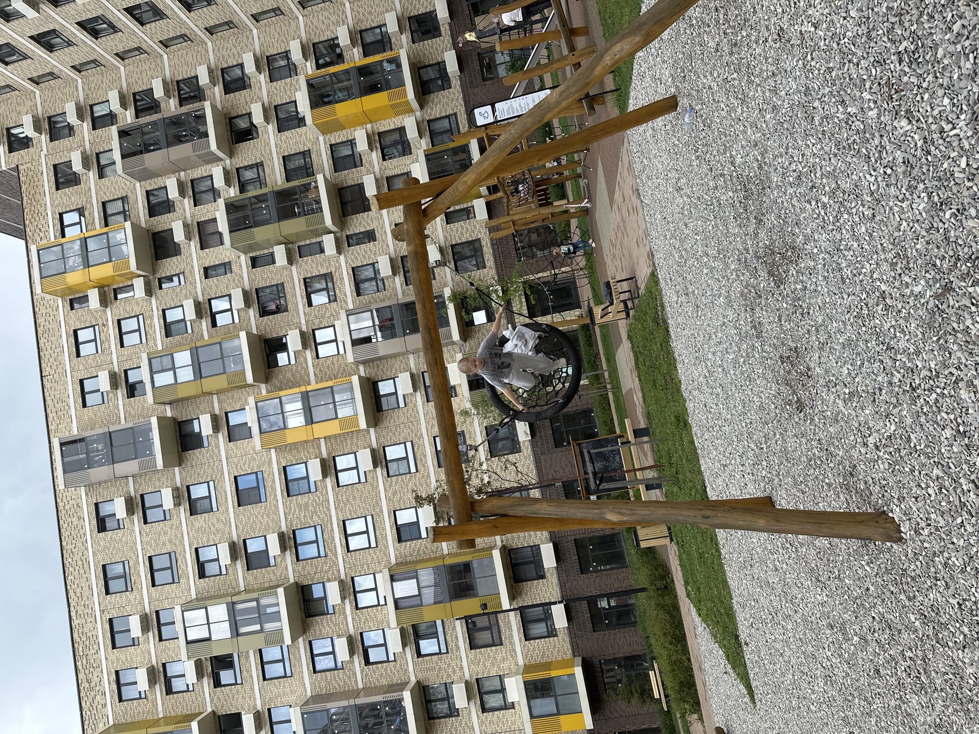 In Znamya Oktyabrya, Russia, a young girl with blonde hair, wearing grey sweatpants and a grey t-shirt, is captured mid-air on a circular net swing. She is positioned on a playground, with a wooden swing set framing the foreground. The swing set's wooden posts are weathered and prominent. Behind the playground, a large, modern apartment building dominates the background. This building features a beige brick facade with numerous windows and balconies, some accented with yellow and grey geometric elements. The ground beneath the swing set is covered in small, light-colored gravel. Other playground equipment, including a climbing structure, is visible further back, along with a few other children. The sky is overcast.