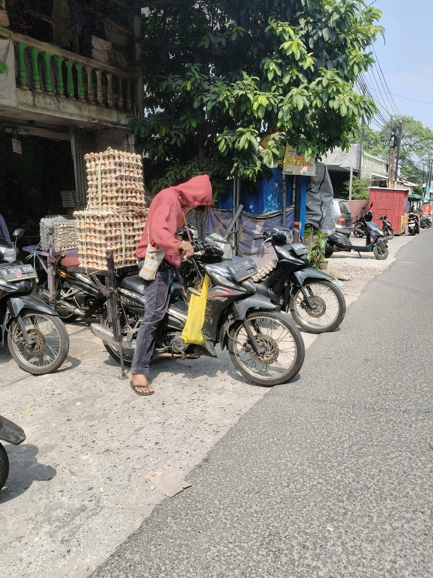 The image depicts a street scene in South Tangerang, Indonesia, with a focus on a person standing next to a motorcycle. The person, wearing a red hooded jacket and dark pants, is on the right side of the frame, with their body partially turned towards the motorcycle. They appear to be adjusting something on the motorcycle or preparing to ride. A yellow bag is hanging from the handlebars, and a small woven bag is slung over their shoulder.
In the background, a stall or shop is visible, featuring a large stack of egg cartons. Several other motorcycles are parked on the side of the road. The environment is sunny, with a clear blue sky and green trees providing some shade. The road surface is asphalt, with a rougher concrete or paved area to the left where the motorcycles are parked. Electrical wires are strung overhead. The overall impression is of a busy, everyday commercial or residential street.