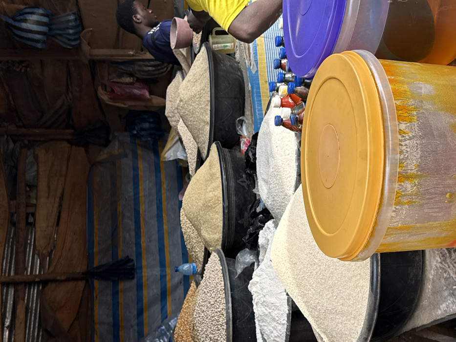 This image depicts an indoor market stall in Ogere, Nigeria. The background consists of corrugated metal sheeting, rough wooden support beams, and a blue and yellow striped tarpaulin. Various bags, including a dark blue striped bag and a red plastic bag, hang from the beams.
The mid-ground is filled with numerous large black plastic basins containing mounded bulk dry goods. These include piles of light-colored pulses (likely beans or peas), light yellow/tan grains (possibly rice or millet), and smaller mounds of similar grains. A small blue plastic water bottle rests on a surface between two mounds of grain.
Two individuals are partially visible. To the left, a male wearing a dark blue shirt with light blue sleeve stripes is seen from the upper chest, looking downwards at a small brown bowl he holds. To the right, only the right shoulder and arm of a person wearing a bright yellow shirt are visible.
In the immediate foreground, a large clear plastic container with an orange-yellow lid shows internal streaks of yellow-orange residue. Adjacent and to its right, a partially visible clear container has a purple lid. A line of small plastic bottles with blue and red caps is arranged behind these foreground containers. Black plastic bags are visible among the basins in the mid-ground.