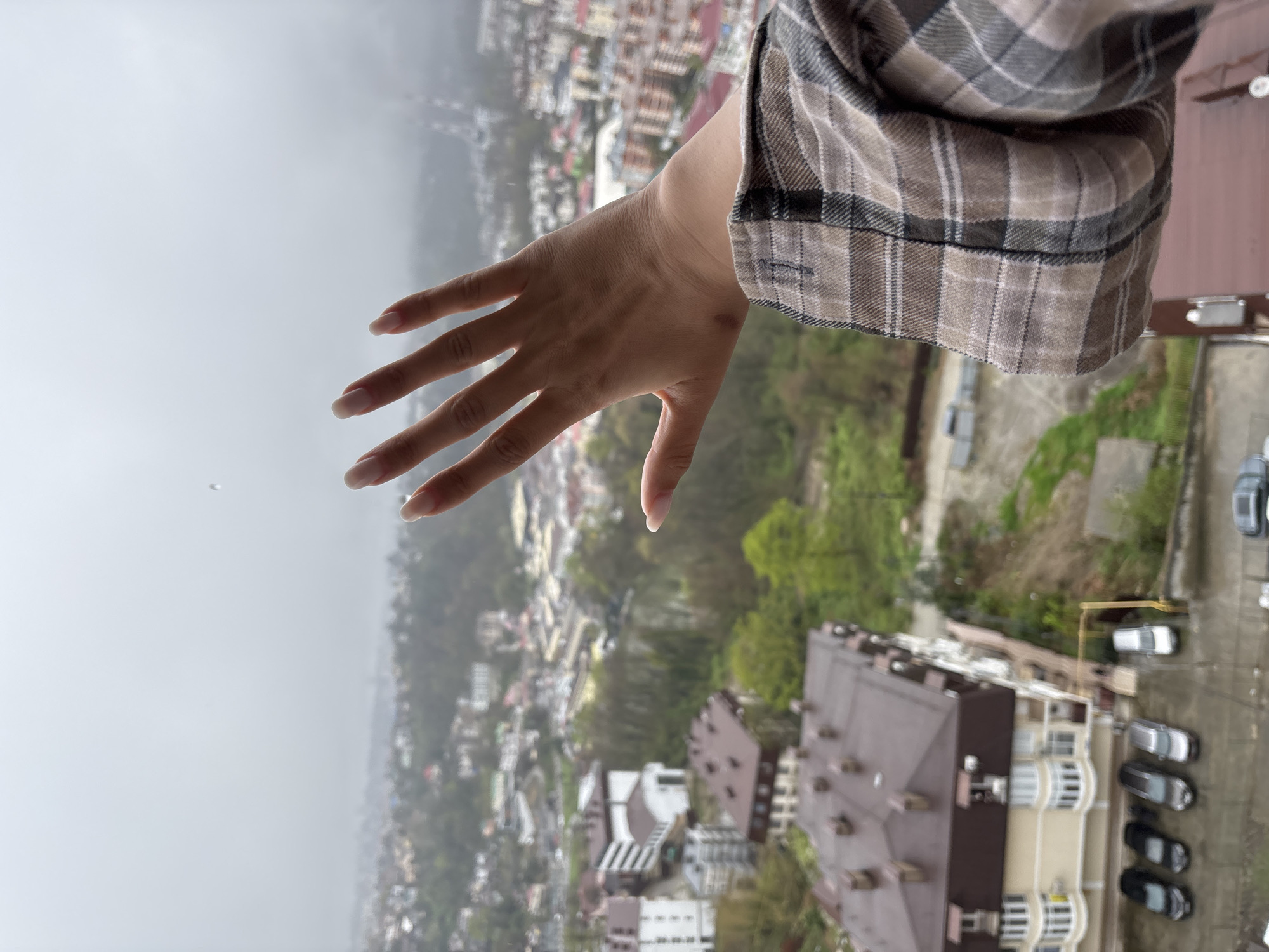 A high-angle, slightly rotated, outdoor shot shows a hand extended over what appears to be a city. The hand, which is of a fair-skinned person, has light pink nails. The sleeve of a plaid shirt is visible at the wrist. The background is a cityscape with buildings, trees, and roads. The environment appears overcast or foggy. In the distance, there is the silhouette of a mountain. Down below is a residential complex. The location may be Sochi, Russia.