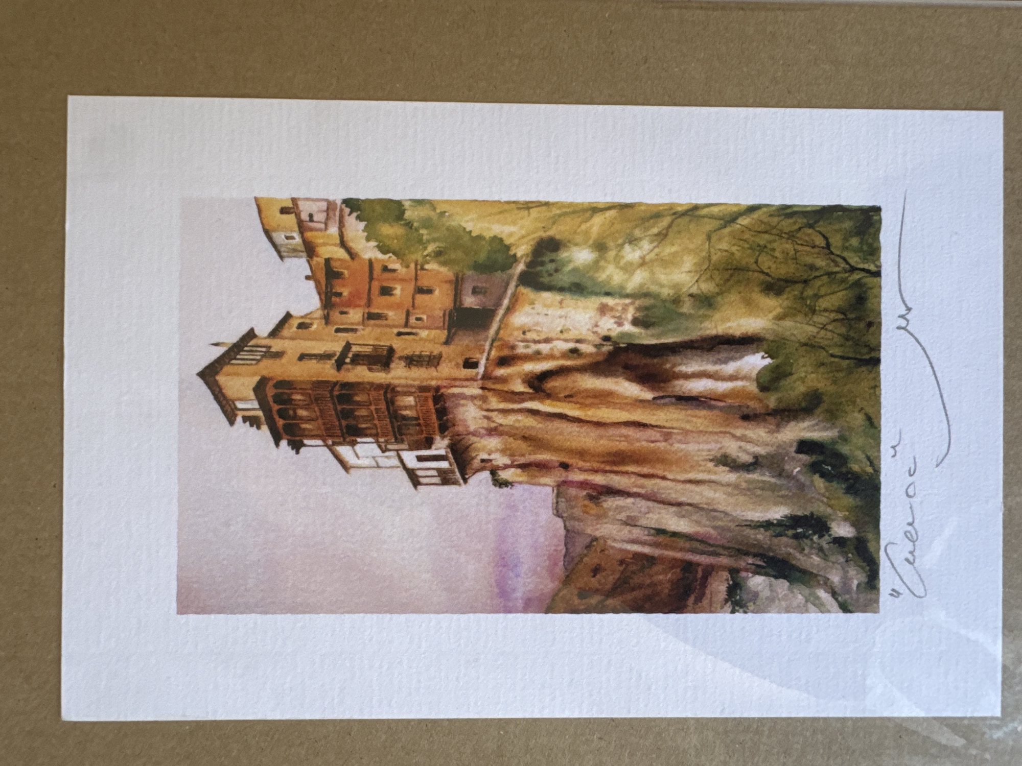 The image is a watercolor painting of the Hanging Houses of Cuenca, a city in the province of Cuenca, Spain, which is located approximately 170 kilometers southeast of Valencia. The painting depicts several multi-story buildings that appear to be constructed directly on the edge of a steep cliff. The architecture features balconies and wooden latticework, with warm, earthy tones dominating the buildings' facades, ranging from yellow to reddish-brown. The cliff itself is rendered with textured brushstrokes, highlighting its rugged, layered geological structure in shades of brown, ochre, and cream.
In the foreground, the base of the cliff is partially obscured by dense foliage, painted in various shades of green and yellow, suggesting trees and undergrowth. The sky above the buildings is a soft, muted pink and grey, indicating either dawn or dusk. Below the painting, on the white matting, there is a handwritten inscription in gray ink. The first part of the inscription appears to be a word enclosed in quotation marks, potentially the title of the artwork or the location it depicts. To the right of this inscription, there is a signature, also in gray ink, consisting of a few curved lines. The overall impression is a picturesque and somewhat dramatic representation of a unique architectural and natural landscape.