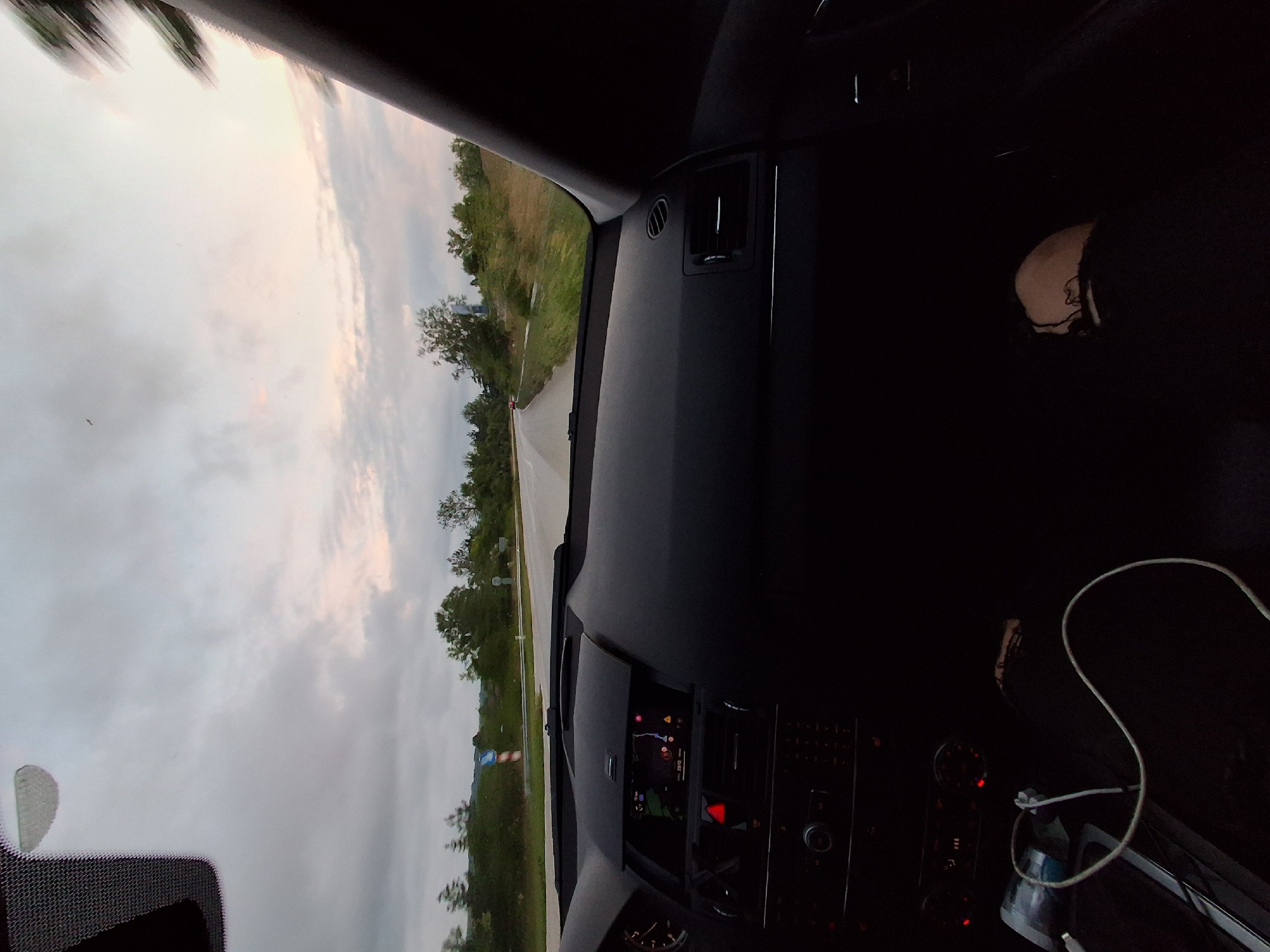 The image, taken from inside a car, provides a view through the front window of a road and the surrounding landscape under an overcast sky. The photograph is oriented vertically, rotated 90 degrees clockwise from a typical horizontal view.
Inside the car, parts of the dark dashboard, including air vents and an active infotainment screen, are visible. The screen displays a navigation map with a blue route line and small numbers, possibly indicating time or distance, such as "2:12" and "2.1". A red triangular symbol, resembling a hazard warning light, is also present on the control panel. A white charging cable is draped over the center console towards the lower right. In the dark passenger area, a faint outline of a person's head and shoulder can be seen, likely a passenger or a reflection of the driver.
Outside the car, a paved road extends into the distance, bordered by grassy areas and scattered trees. A small, unreadable blue and white road sign stands on the right side of the road. The sky is predominantly overcast with grey clouds, but a subtle lighter, yellowish-pink hue near the horizon suggests the time of day is late afternoon or early evening, likely dusk. The setting appears to be a rural or semi-rural environment, consistent with the specified location of Novo Selo, Bulgaria.
The activity depicted is travel by car, indicated by the active navigation system.