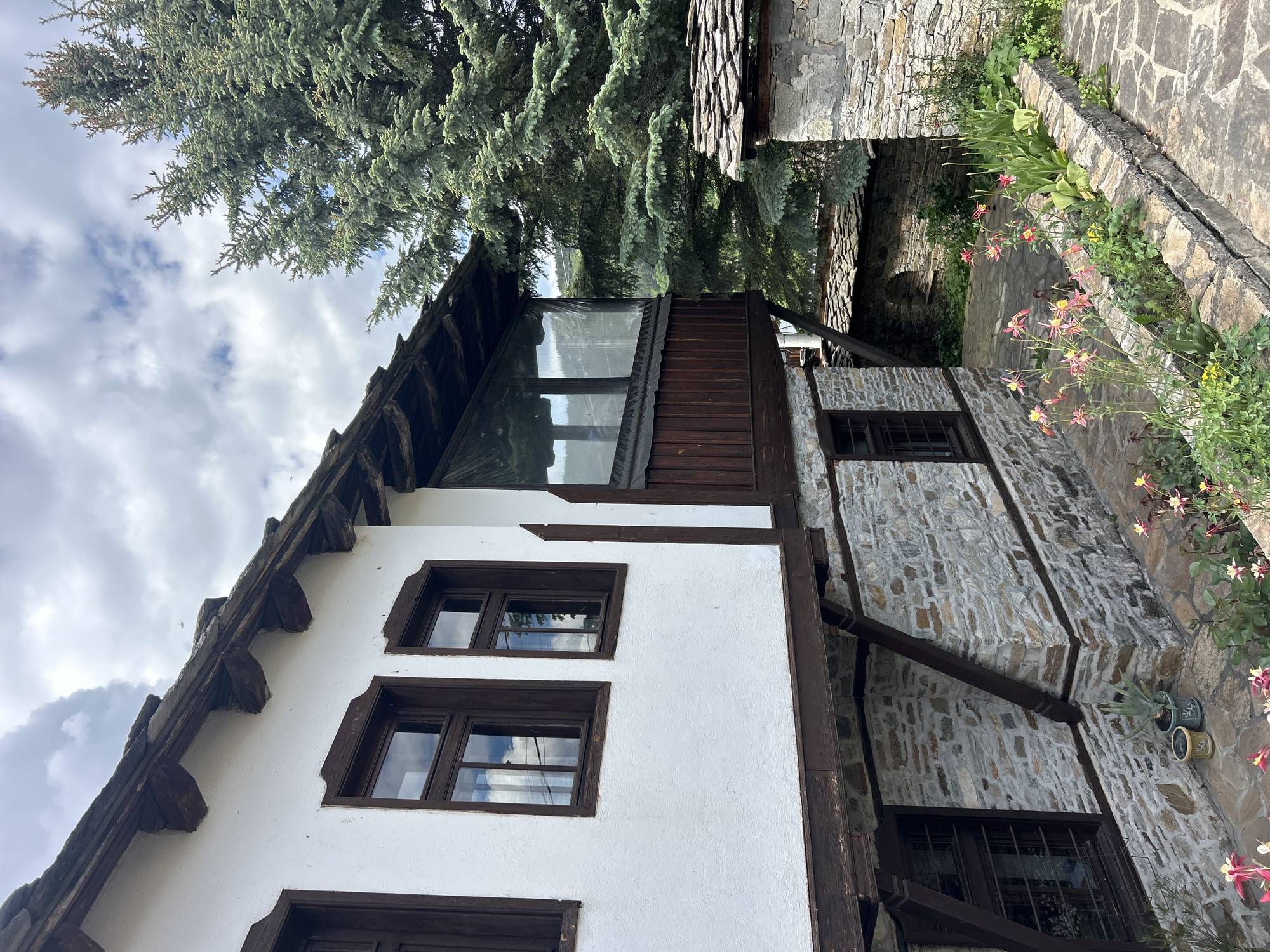 The image depicts a traditional Bulgarian house in the village of Dolen, Bulgaria, characterized by its white plastered walls and dark wooden framing elements. The house has several windows with dark frames, some reflecting the sky. The roof is tiled with a visible overhang and decorative wooden supports. A small enclosed balcony or veranda with a translucent window is present. The house is partly constructed with stone masonry on one side, and a stone pathway with flowering plants runs along the edge. A large tree looms over the house, partially obscuring the sky. The sky is cloudy. The image is taken from a slightly low angle.