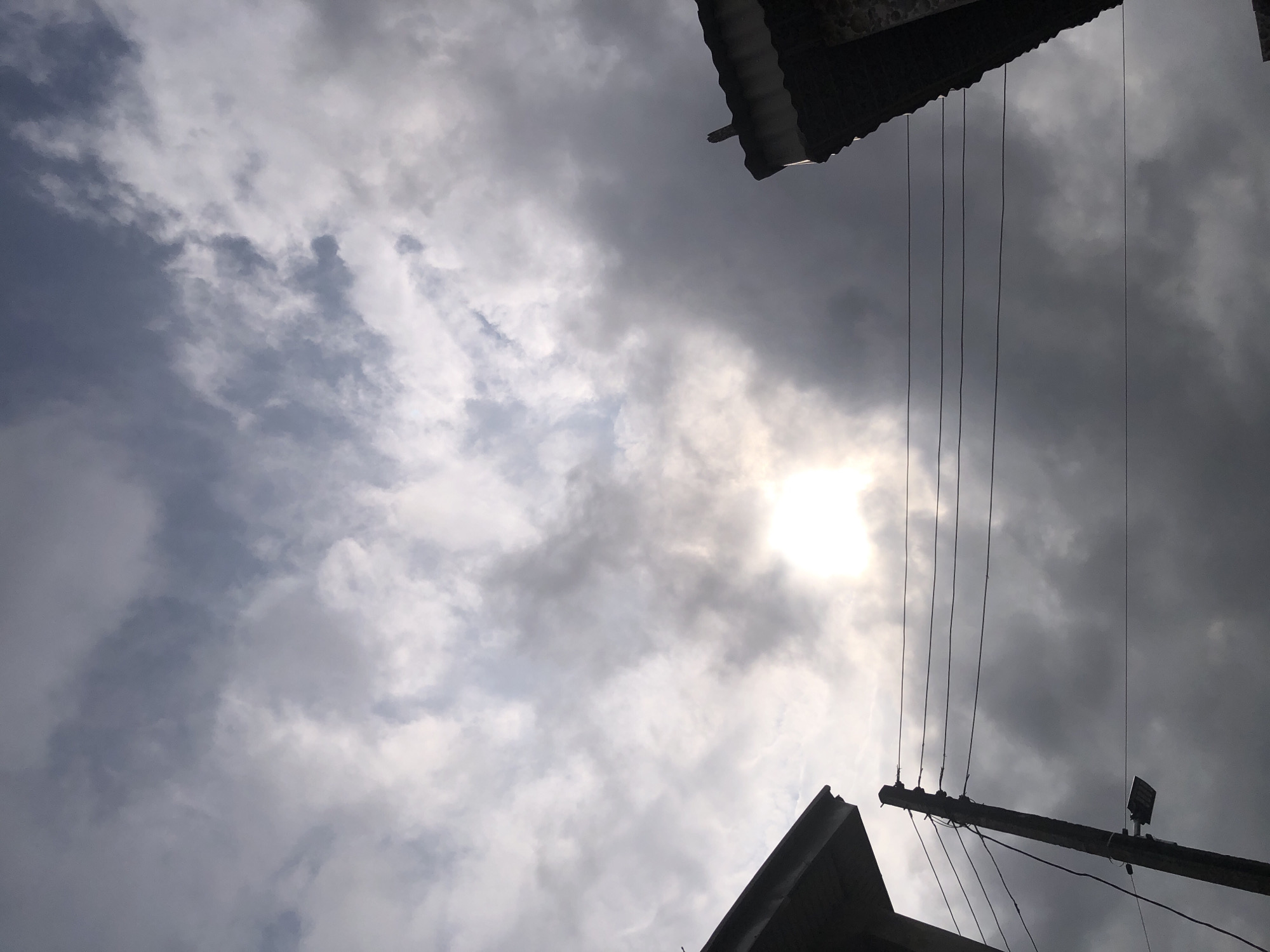 The image depicts a cloudy sky over Ado, Nigeria. The sun is visible, partially obscured by the clouds, creating a bright halo effect. In the foreground, the edge of a building with a tiled roof is visible on the right, and another building's corner is seen on the left. A utility pole stands centrally, with multiple wires stretching horizontally across the frame. A street lamp is attached to the pole, and another wire is visible near it. The overall atmosphere is overcast, suggesting the possibility of rain or a storm. The scene appears to be an outdoor urban environment, likely a residential or commercial area.