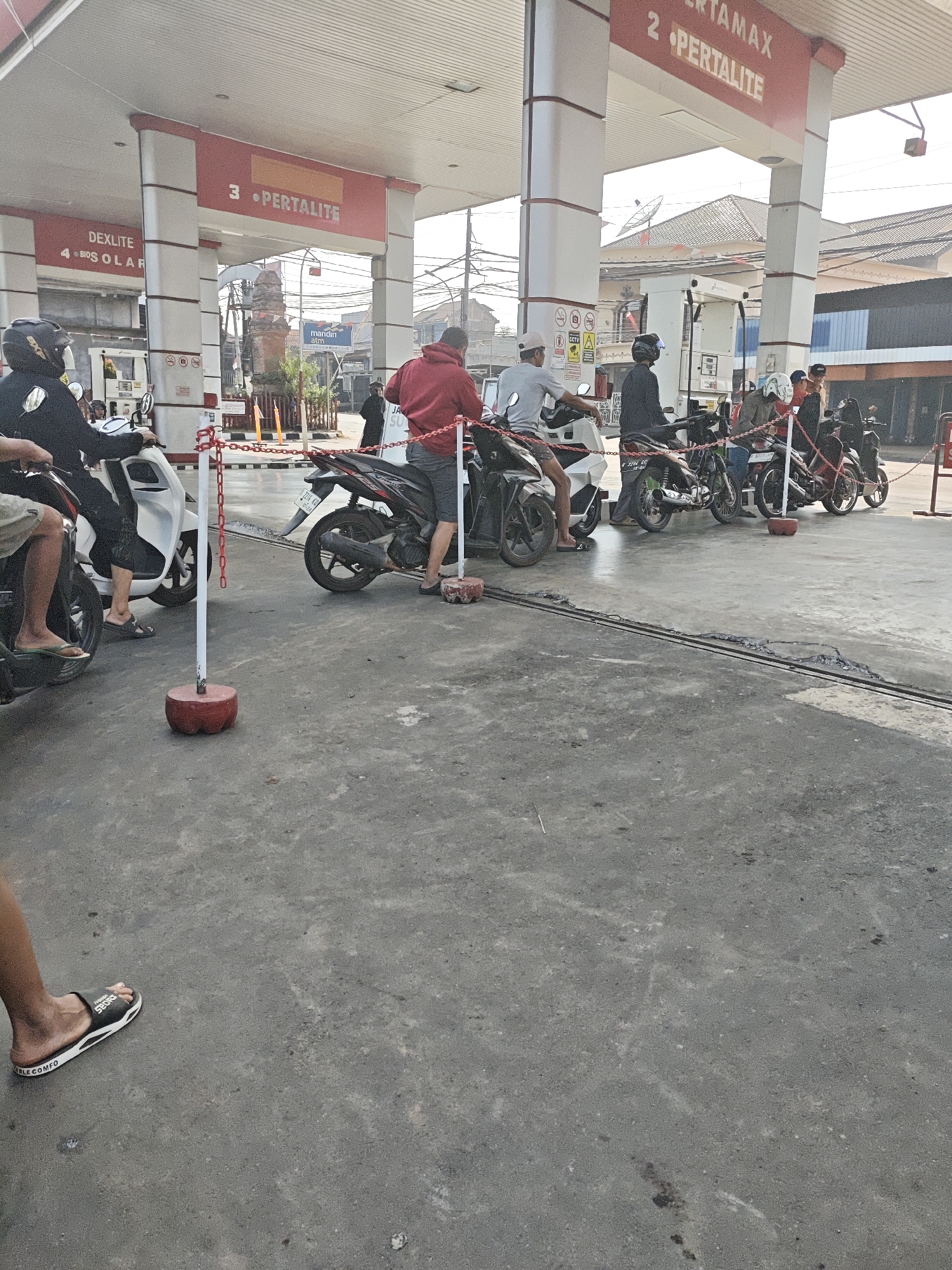 The image shows a queue of motorcycles at a gas station in Ngabul, Indonesia. The gas station has red and white pillars with signs indicating fuel types like "PERTAMAX" and "PERTALITE". Several people are waiting on their motorcycles, some wearing helmets. The ground is paved with asphalt. In the background, there are buildings and some vegetation. The scene suggests a normal day at a busy gas station.