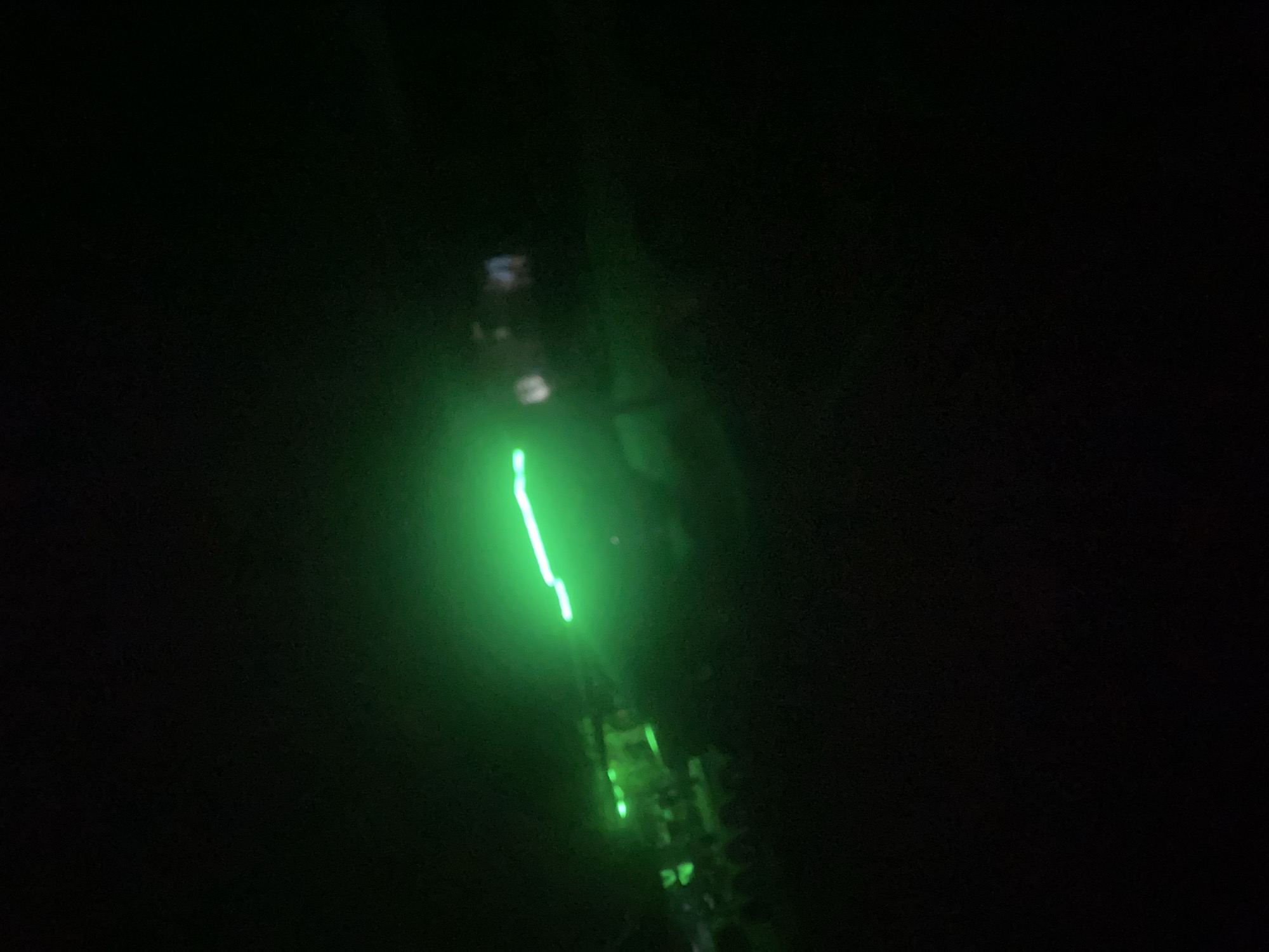 The image shows a dark outdoor scene with a prominent green light source. The light emanates from what appears to be a long, thin object, possibly a laser pointer or a string of lights, extending from the left side of the frame towards the right. The light beam is bright and somewhat blurred, indicating movement or a long exposure. In the background, there are faint shapes of what could be structures or trees, also illuminated by the green light. The overall impression is of a nighttime setting. The provided image does not contain enough information to determine the location within the city of Howayi, Nigeria.