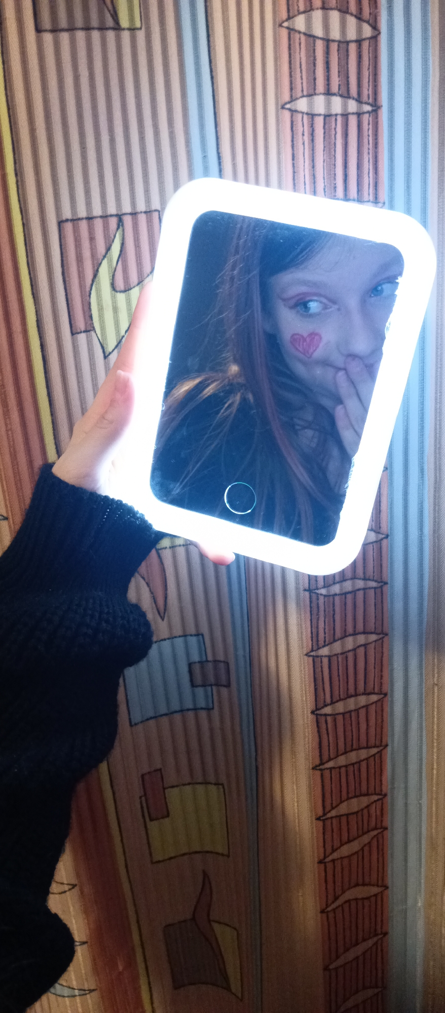 A hand holds a light-up mirror reflecting a person with a heart drawn on their cheek. The person is glancing to the side with their hand covering their mouth. The background is a striped fabric with abstract shapes.
