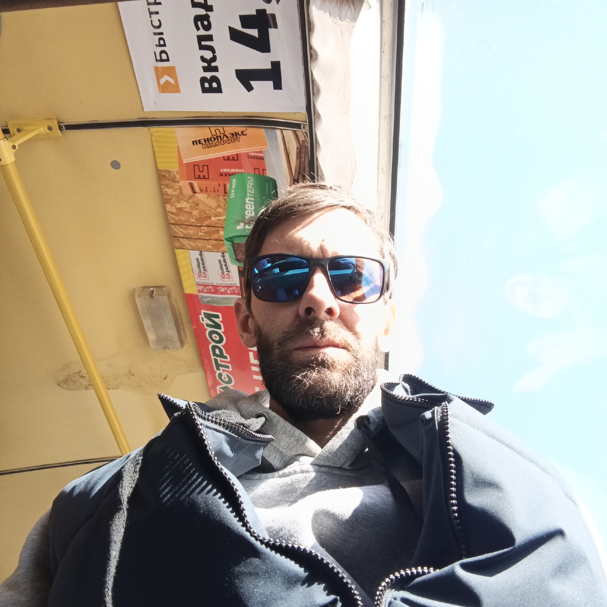 A man is captured in a close-up, low-angle selfie, positioned in the foreground of the image. He has a full beard and is wearing dark rectangular sunglasses with blue reflective lenses, which reflect the bright sky and clouds. He is dressed in a grey hooded sweatshirt under a dark blue zip-up jacket, which is partially unzipped. His expression is neutral.
The setting appears to be the interior of a vehicle, possibly public transport like a bus or tram, or a commercial establishment, given the advertisements visible on the yellow ceiling and upper walls. A bright window to the right reveals a clear, blue sky, indicating a sunny daytime. A yellow metal bar runs across the top left of the frame.
Various advertisements and product labels, mostly in Russian, are visible in the background. Notable text includes:
- "> Быстр" (Fast)
- "Вклад" (Contribution/Deposit, possibly part of a larger phrase)
- "14" (Number 14)
- "ПЕНОПЛЭКС КОМФОРТ" (PENOPLEKS COMFORT), a brand for insulation materials.
- "greenTERM", another potential brand name related to construction or insulation.
- "Изоляция Базальтова" (Basalt Insulation).
- "0,6 м² внутри" (0.6 m² inside).
- "НПО Строй" (NPO Stroy), likely a construction-related entity.
The lighting is strong, casting shadows from the man onto his clothing and highlighting parts of his face. The overall impression is a casual, everyday scene in Chaikovsky, Russia.