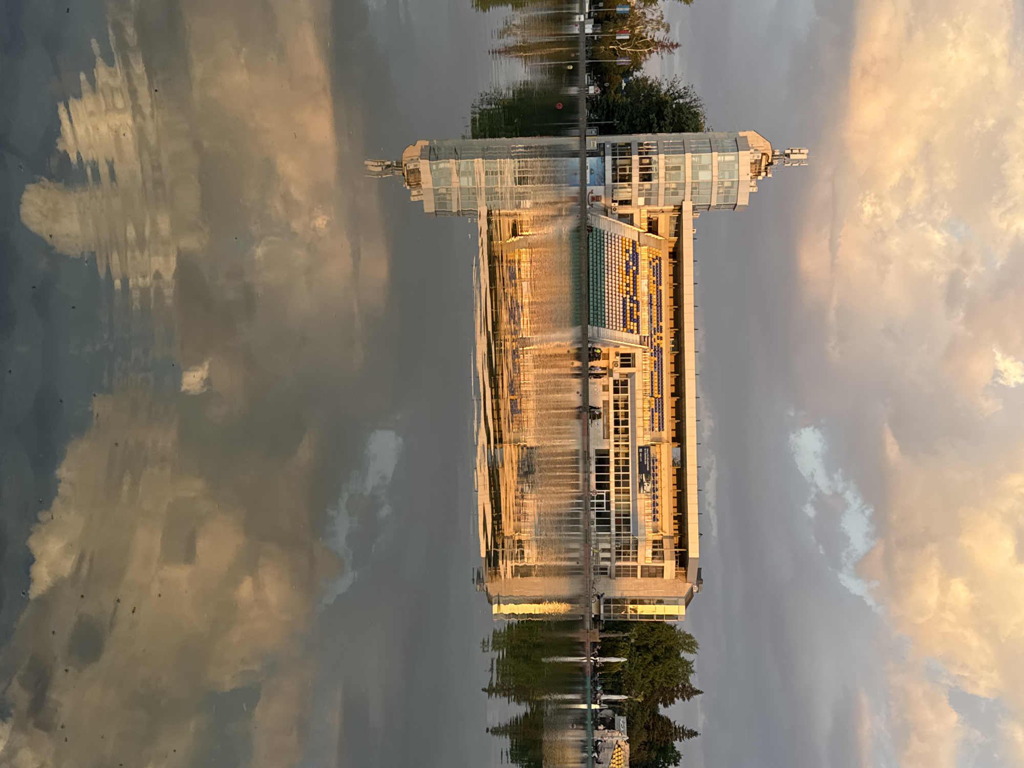 The image shows the reflection of the Plovdiv Rowing Canal building and the surrounding sky in the water. The building appears elongated vertically due to the reflection. The sky contains a mix of gray and golden-hued clouds, indicative of either sunrise or sunset. The building is a modern structure with a partially visible seating area colored in blue, green, yellow, and white. Some trees are visible in the foreground at the bottom of the image. The overall lighting suggests either early morning or late afternoon conditions.