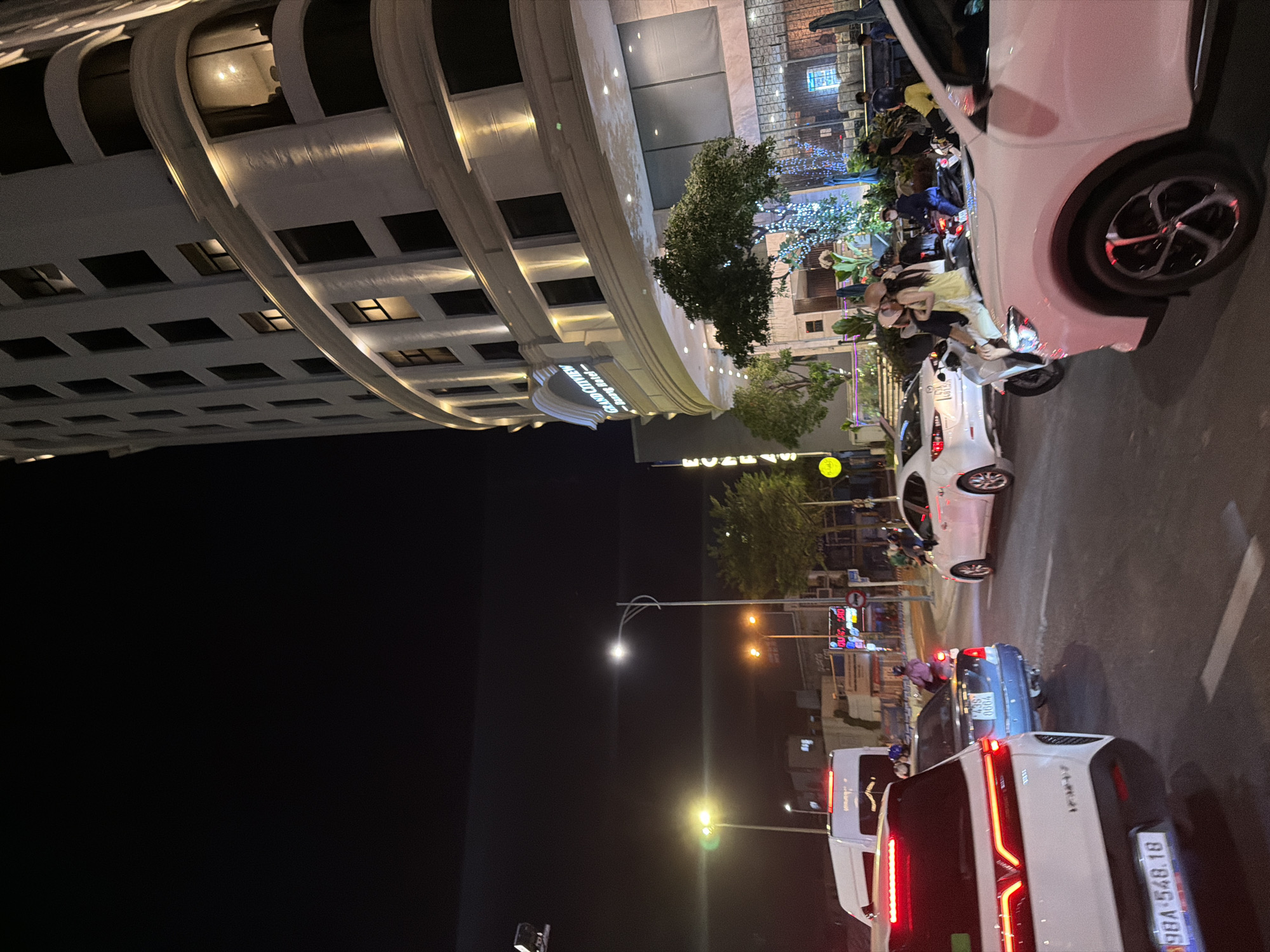 This is a night-time, street-level view of a busy intersection in Da Nang, Vietnam. The scene is dominated by vehicles, including cars and numerous motorcycles. On the left, a modern building with curved architectural elements and illuminated windows looms over the street. The building appears to be a hotel or commercial establishment, with a sign that reads "Grand Plaza Hotel."
Several white cars are visible, including one in the foreground with its taillights illuminated, displaying the license plate "98A-548.18." A blue car is also parked in traffic. A multitude of motorcycles, some carrying multiple passengers, are interspersed among the cars, highlighting the common mode of transportation in the area. People are seen on the motorcycles, some wearing helmets.
Streetlights and the lights from buildings and vehicles illuminate the scene, creating a vibrant and dynamic atmosphere. The time of day is clearly night. The weather appears to be clear. The presence of traffic lights and the general hustle and bustle suggest a typical evening in a Vietnamese city. There are some trees and decorative lights visible in the background, adding to the urban ambiance. The overall impression is one of urban activity and movement at night.