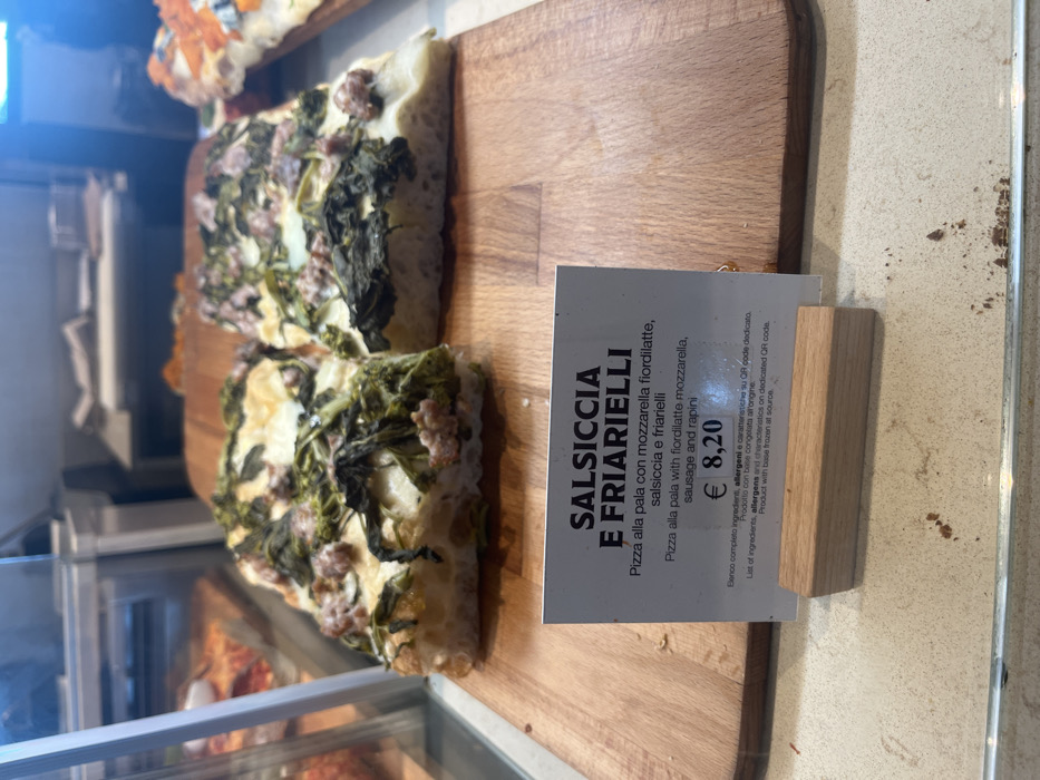 The image captures a close-up view of two rectangular slices of "Salsiccia e Friarielli" pizza displayed on a light-colored wooden cutting board, likely within a glass food display case in a bakery, pizzeria, or deli in Dorno, Italy. The pizza slices feature a thick, airy crust, topped with mozzarella fiordilatte cheese, crumbled sausage, and cooked rapini (broccoli rabe).
To the right of the pizza, a white price and description tag is prominently displayed, held upright by a small wooden stand. The tag provides details in both Italian and English. The main title reads "SALSICCIA E FRIARIELLI." Below this, the Italian description states "Pizza alla pala con mozzarella fiordilatte, salsiccia e friarielli," followed by the English translation: "Pizza alla pala with fiordilatte mozzarella, sausage and rapini." The price for this pizza is clearly marked as "€ 8,20."
Further text on the tag indicates, "Elenco completo ingredienti, allergeni e caratteristiche su QR code dedicato" (List of ingredients, allergens and characteristics on dedicated QR code) and "Prodotto con base congelata all'origine" (Product with base frozen at source). The setting appears to be indoors with artificial lighting, showcasing the prepared food for sale. Part of another pizza or baked good is visible in the upper left background, suggesting a variety of items are available.