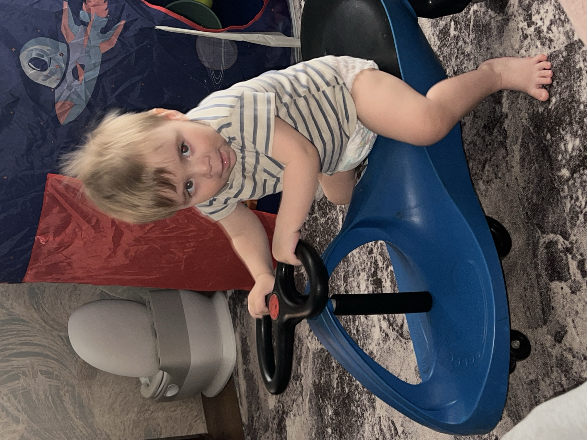 A child, approximately one year old, is lying on a blue toy car on a patterned carpet. The child is wearing a striped shirt and a diaper. The child's right leg is extended towards the upper right of the frame. The child is gripping the black steering wheel of the toy car. Behind the child is a play tent with a space-themed design. Next to the tent, a gray and white air purifier. The photograph is taken from an overhead perspective. The location is Noviy, Russia.
