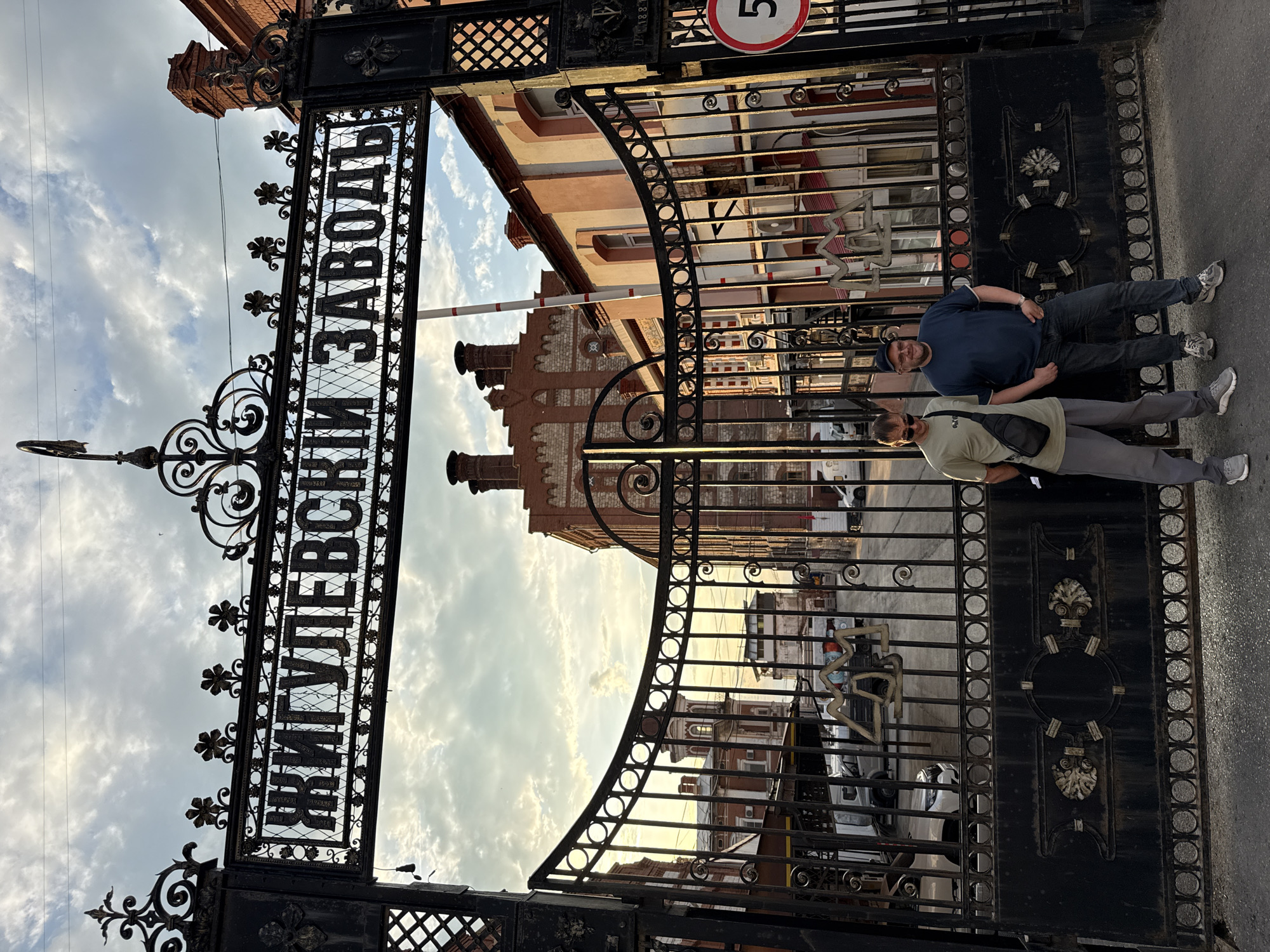 Two men stand in front of a large, ornate black iron gate. The gate has decorative scrollwork and features the Cyrillic text "ЖИГУЛЕВСКИ ЗАВОДЪ" (Zhigulevsky Zavod) inscribed across its upper portion. Beyond the gate, a courtyard is visible with several red brick industrial buildings with white trim, characteristic of turn-of-the-century architecture. Sunlight illuminates the scene from the right, casting long shadows. The sky above is partly cloudy with a mix of light and dark clouds. The man on the left wears a light olive t-shirt, grey pants, and sunglasses, with a black crossbody bag. The man on the right wears a dark blue t-shirt, dark jeans, and a baseball cap. The gate itself is a significant architectural feature, suggesting a historical or industrial site. The overall impression is that of an entrance to a historic factory or brewery. The inscription on the gate and the architectural style strongly suggest the location is the Zhigulevsky Brewery in Samara, Russia.