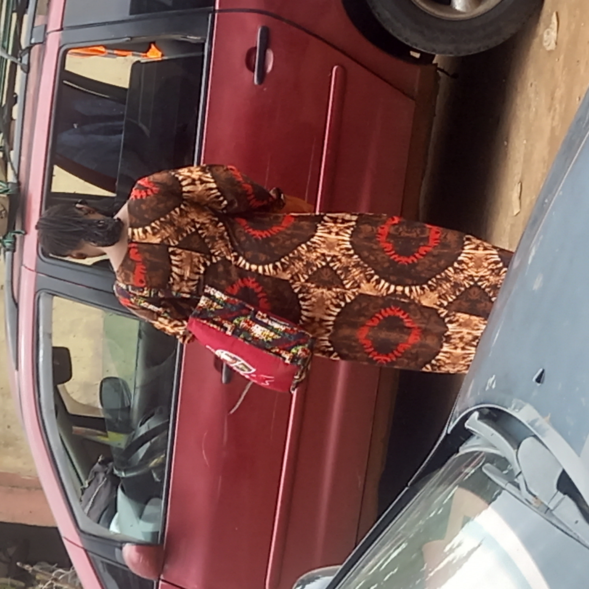 A woman stands next to two cars in Sardauna, Nigeria. She is wearing a brown and red patterned dress and carrying a red handbag. The car to her left is red, and the car to her right is gray. The environment appears to be a street or parking area.