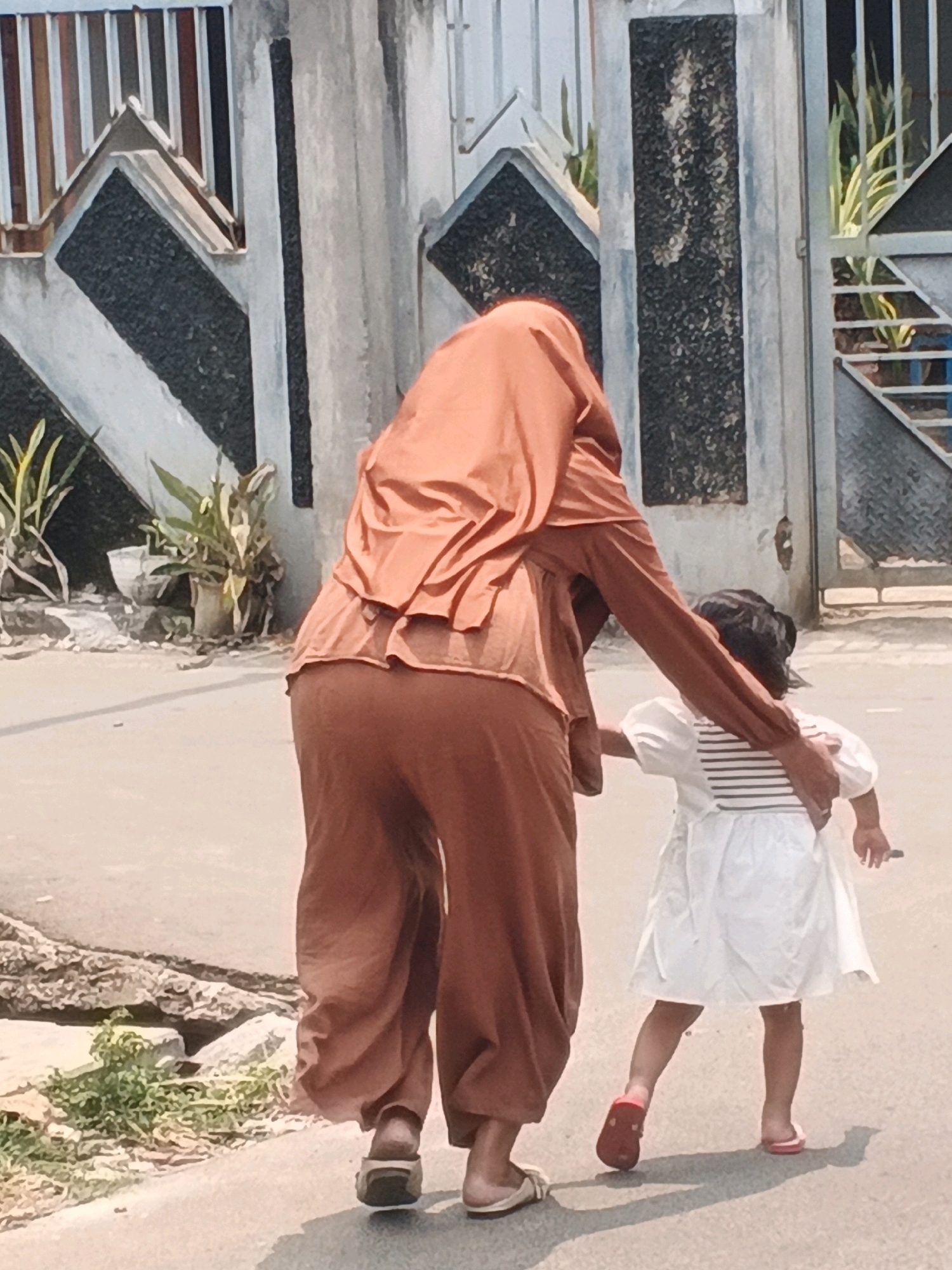 Two individuals are walking on a paved surface, likely a road, in Kadu, Indonesia. The person in the foreground, from the rear, is wearing an orange headscarf and loose orange pants. Their right arm is around the shoulders of a child. The child in front is wearing a white dress with horizontal black stripes on the chest. The child is wearing red sandals on their left foot and pink sandals on their right. The environment appears to be outdoors, with a wall and fence in the background. The wall is made of concrete with dark textured panels and metallic elements, possibly part of a gate or decorative feature. Plants are visible growing near the wall. The lighting suggests it is daytime.