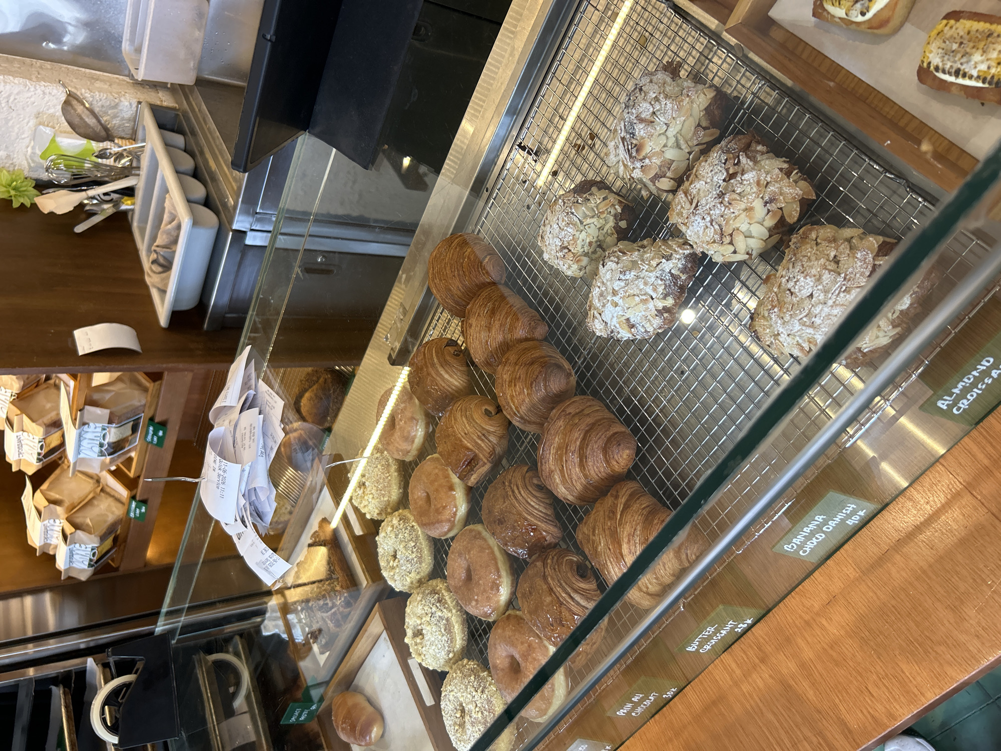 This image shows a display case filled with baked goods, likely in a bakery or cafe in North Kuta, Indonesia. The display case is made of glass and metal, and it is filled with a variety of pastries, including croissants, donuts, and danishes. Some of the pastries are dusted with powdered sugar and almonds, while others are glazed. There are also some receipts piled on top of the display case. In the background, there are shelves stocked with more baked goods in paper bags. The overall impression is one of a well-stocked and inviting bakery.