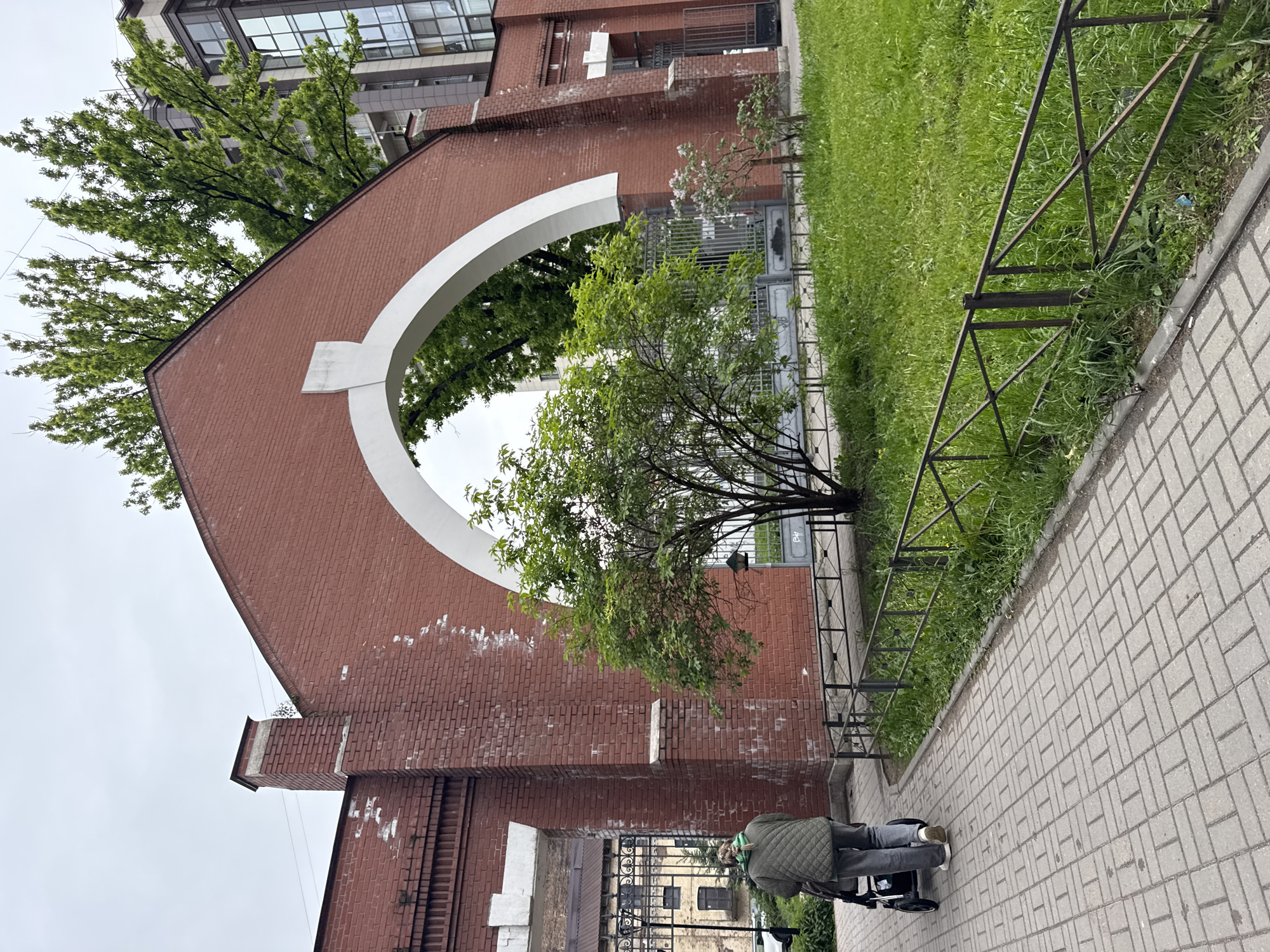 The image shows a person with a stroller walking on a brick-paved sidewalk in front of a large brick archway structure with a white circular opening. The archway is part of a building with a modern extension on top. Green trees and grass are visible behind the arch and on the side of the walkway. A metal fence runs alongside the walkway, separating it from the grassy area. The archway structure appears to be located in Saint Petersburg, Russia, based on the architectural style.