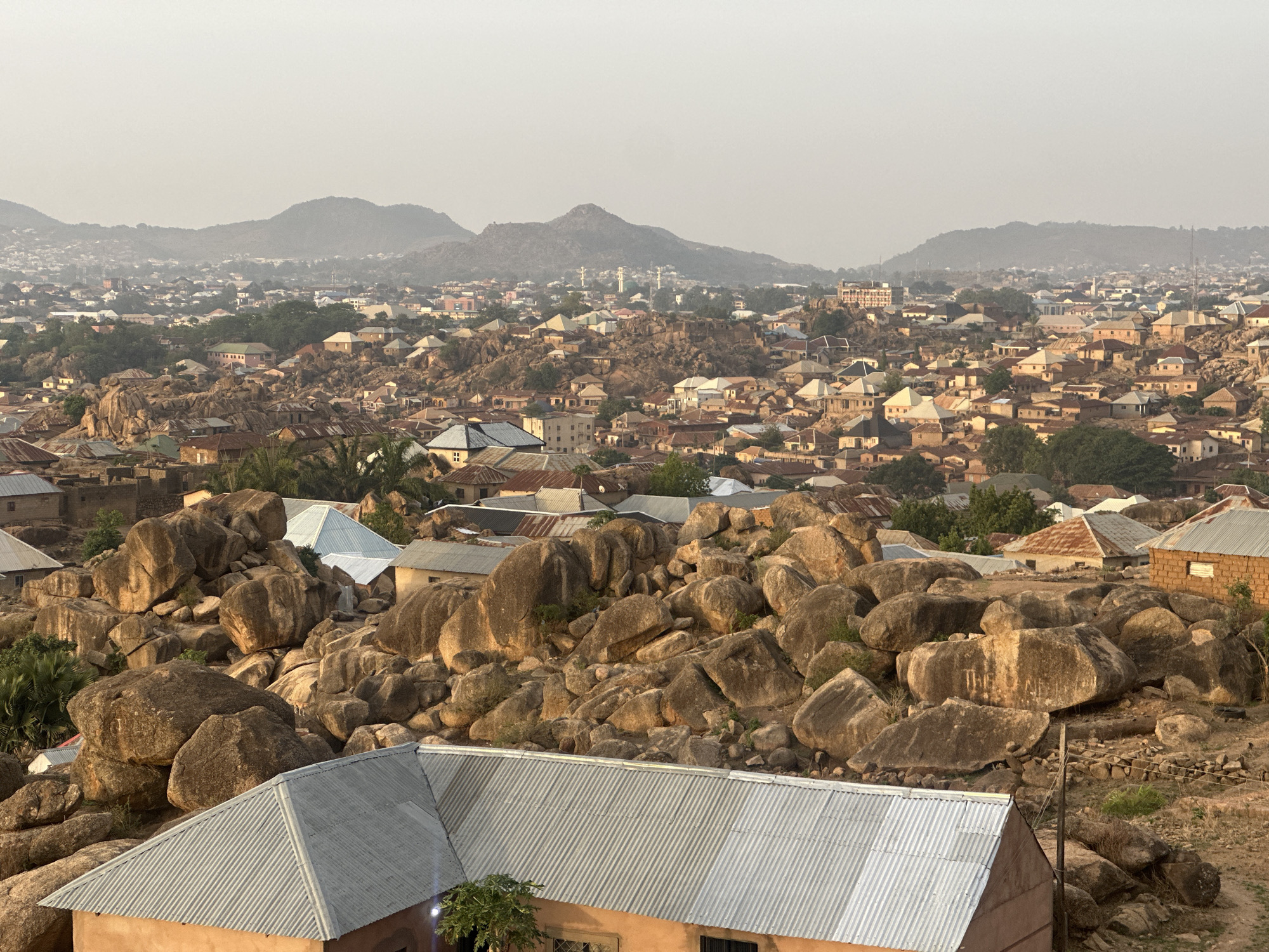 The image depicts an elevated view of a residential area in Jos, Nigeria. The scene is characterized by a landscape interspersed with large, rounded boulders amidst buildings. The structures vary in size and design, many with corrugated metal roofs. The foreground features a building with a gray, corrugated metal roof. In the distance, hazy mountains rise beyond the densely populated urban area. The overall light suggests either dawn or dusk, with soft shadows cast across the scene.