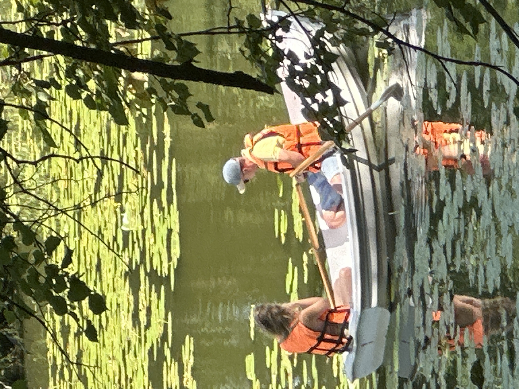 Two people are in a rowboat on a lake covered in lily pads. The person in the front of the boat, facing away from the camera, is wearing an orange life vest and has their hand to their head. The person in the back of the boat, facing the camera, is wearing a yellow shirt and an orange life vest and is holding an oar. The boat is white with a gray stripe. The water is green and reflects the boat and people. The background is a mix of green foliage and trees. The scene appears to be in Ostafievo, Russia, a location known for its historic estate and parkland which includes a lake suitable for boating.