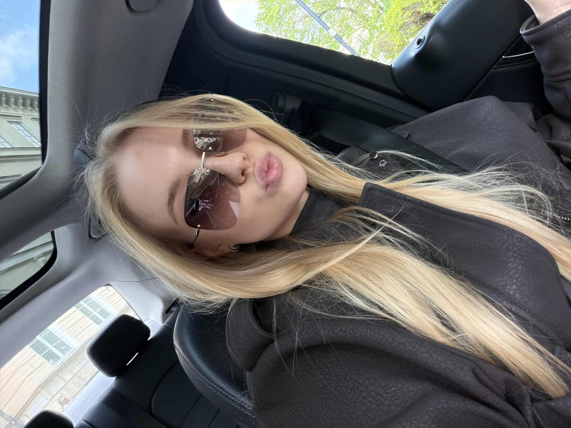 A woman with blonde hair and sunglasses is sitting in a car. She is wearing a black jacket. The interior of the car is black leather. Outside the window, there is a building and some trees. The location appears to be a city street, but the specific city is unknown. The woman is looking at the camera with pursed lips.
