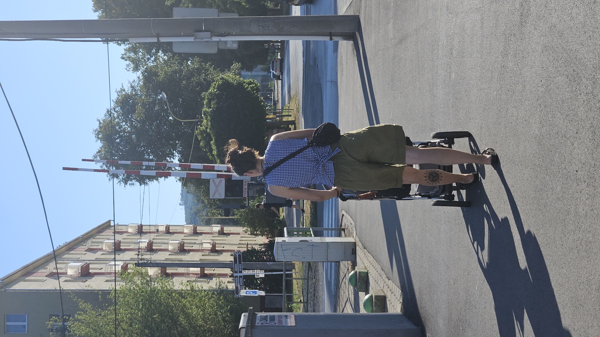 A woman with dark, tied-back hair is walking away from the camera, pushing a stroller. She is wearing a blue and white checkered sleeveless shirt with a tie in the back and loose olive green shorts. She has a dark, rounded bag slung across her body. A tattoo of a circular, sun-like pattern is visible on her left calf. She is wearing dark sandals. The stroller is dark-colored with two large rear wheels and two smaller front wheels. The woman is walking on an asphalt path.
In the background, there is a multi-story building with a light-colored facade and balconies with red railings. To the left of the building, a grey trash receptacle with a sticker is visible. Green, rounded bollards are lined up along the edge of the path. Further in the background, there are trees and a railroad crossing with red and white striped barriers. A sign with a red 'X' symbol is positioned below the barriers, and a smaller sign below that reads "POZOR VLAK," which translates to "CAUTION TRAIN" in Slovak. A street with cars is visible beyond the railroad crossing. The sky is clear and blue, and the sun casts long shadows on the ground. The scene appears to be in a residential area in the city of Prešov, Slovakia.
