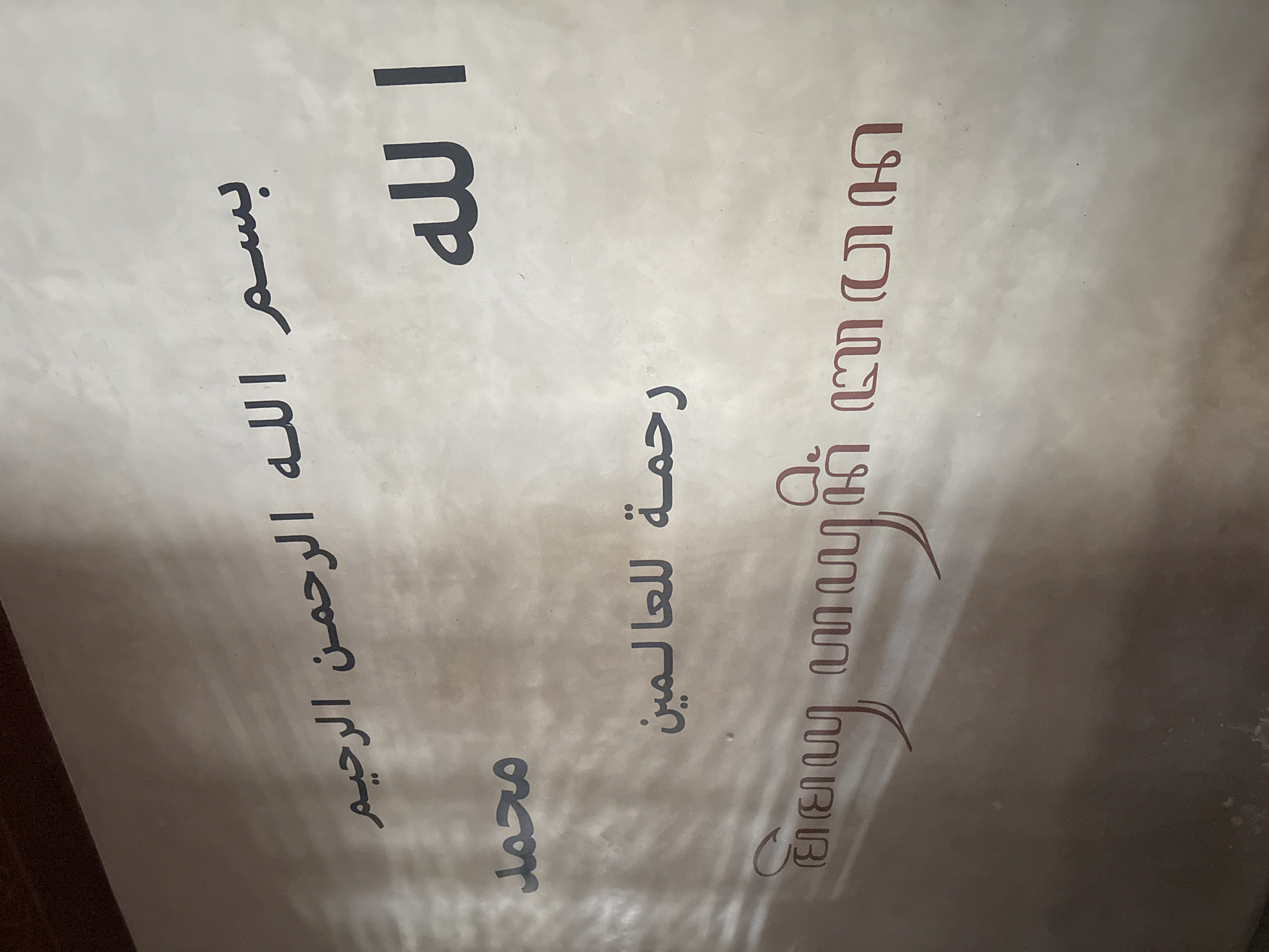 The image displays a vertical panel of text, presented against a light, off-white background with subtle reflections, suggesting it is either a framed piece under glass or a laminated print. The text is oriented vertically when the image is rotated 90 degrees counter-clockwise.
On the left side, rendered in clear, dark black Arabic script, are four lines of religious phrases. From top to bottom, these read:
1. **الله** (Allah, meaning God)
2. **بسم الله الرحمن الرحيم** (Bismillah al-Rahman al-Rahim, meaning "In the name of Allah, the Most Gracious, the Most Merciful")
3. **رحمة للعالمين** (Rahmatan lil-alamin, meaning "A mercy to the worlds" or "A mercy to mankind")
4. **محمد** (Muhammad)
On the right side of the panel, a highly stylized and ornate script, rendered in a warm brown color, is visible. This script is visually complex with intertwined and looping characters, reminiscent of traditional calligraphic styles or indigenous scripts adapted for Islamic expressions, potentially in the Indonesian context. While precise transliteration is challenging due to its intricate design, its presence alongside the explicit Arabic religious phrases strongly suggests it conveys complementary Islamic declarations, possibly a decorative rendering of the Shahada (the Islamic creed).
The overall subject of the media file is Islamic religious calligraphy or inscription, likely functioning as a decorative or devotional item in a setting like a home or mosque in Kalijaten, Indonesia. There are no people, explicit activities, or specific objects other than the textual panel. The background is simple and uncluttered, and no time of day or weather details are discernible.
