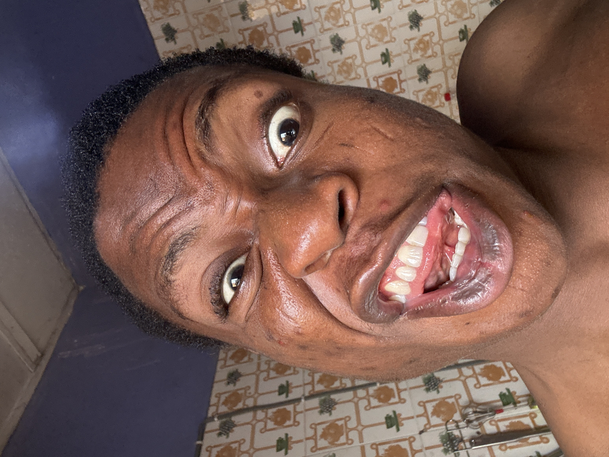 A close-up, angled self-portrait of a dark-skinned individual with black hair. The person is facing the camera with a contorted facial expression, mouth open revealing teeth and tongue. The eyes are wide and directed towards the camera. The skin appears shiny or possibly sweaty. The background is partially visible and shows a tiled wall with an repeating geometric pattern and a blue section. The scene is in Unknown, Unknown.