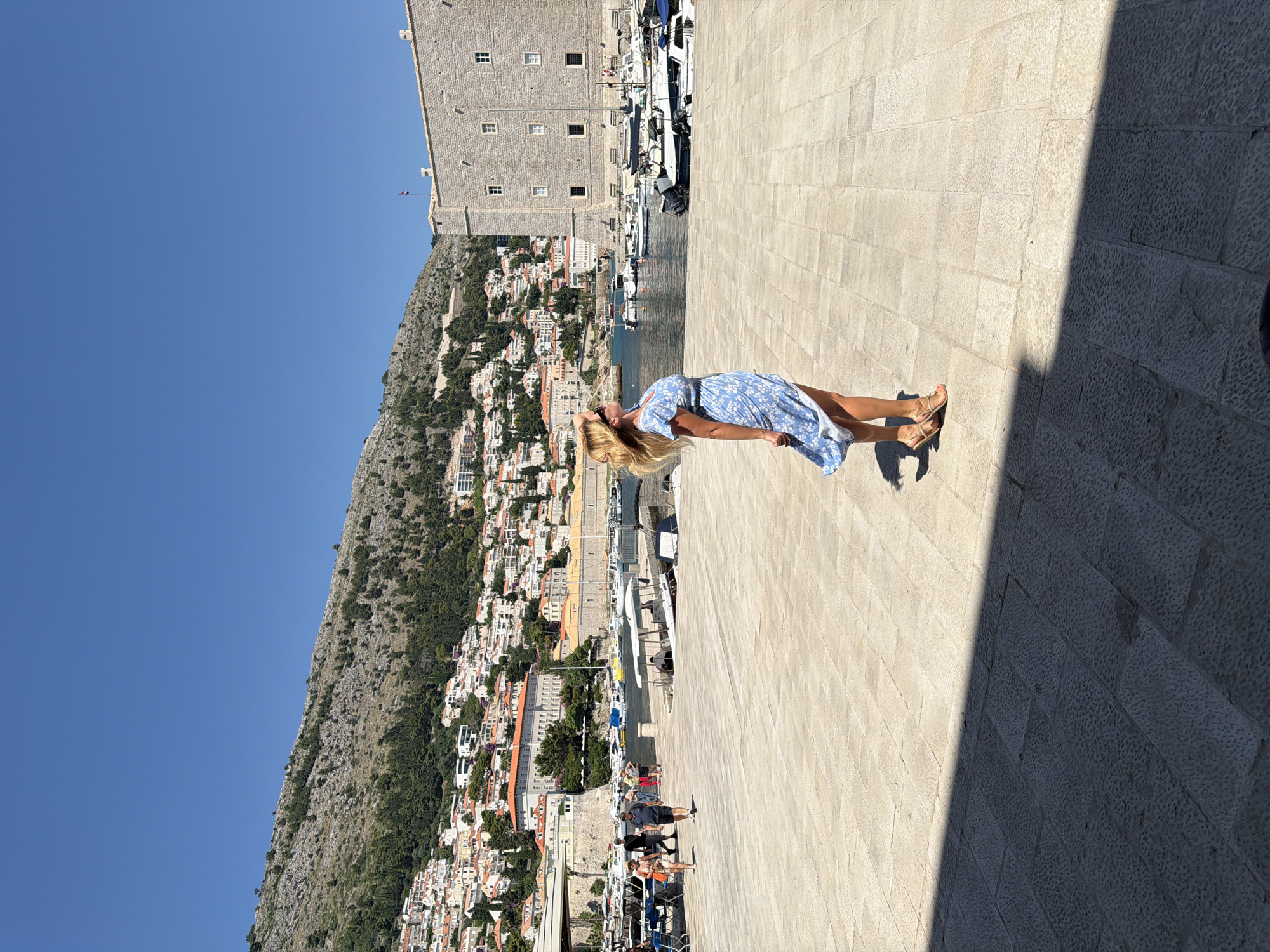 A blonde woman in a blue and white floral dress stands on a stone plaza in Dubrovnik, Croatia, with her head tilted back and hair blowing in the wind. She is facing away from the camera, looking towards a body of water filled with boats. Behind her, a fortified stone building, likely part of the city walls, is visible. In the background, a hillside covered with white buildings and green trees slopes upward, with a clear blue sky overhead. Other people are visible walking in the distance on the plaza and near the boats. The scene appears to be a sunny day in a historic coastal city.