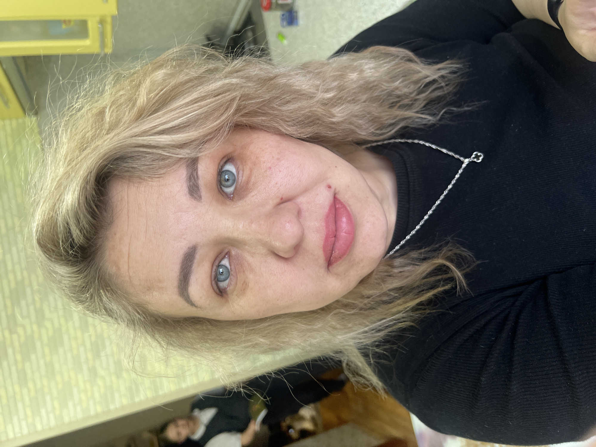 A Caucasian woman with blonde wavy hair, light blue eyes, and a silver necklace with a clover-shaped pendant is facing the camera. She is wearing a black sweater. Red spots appear on her face.
The background features a partially visible yellow cabinet. A person and objects are visible in the lower left corner of the image, in what appears to be a room.
No information confirms this location is in the city of Chaikovsky, Russia.