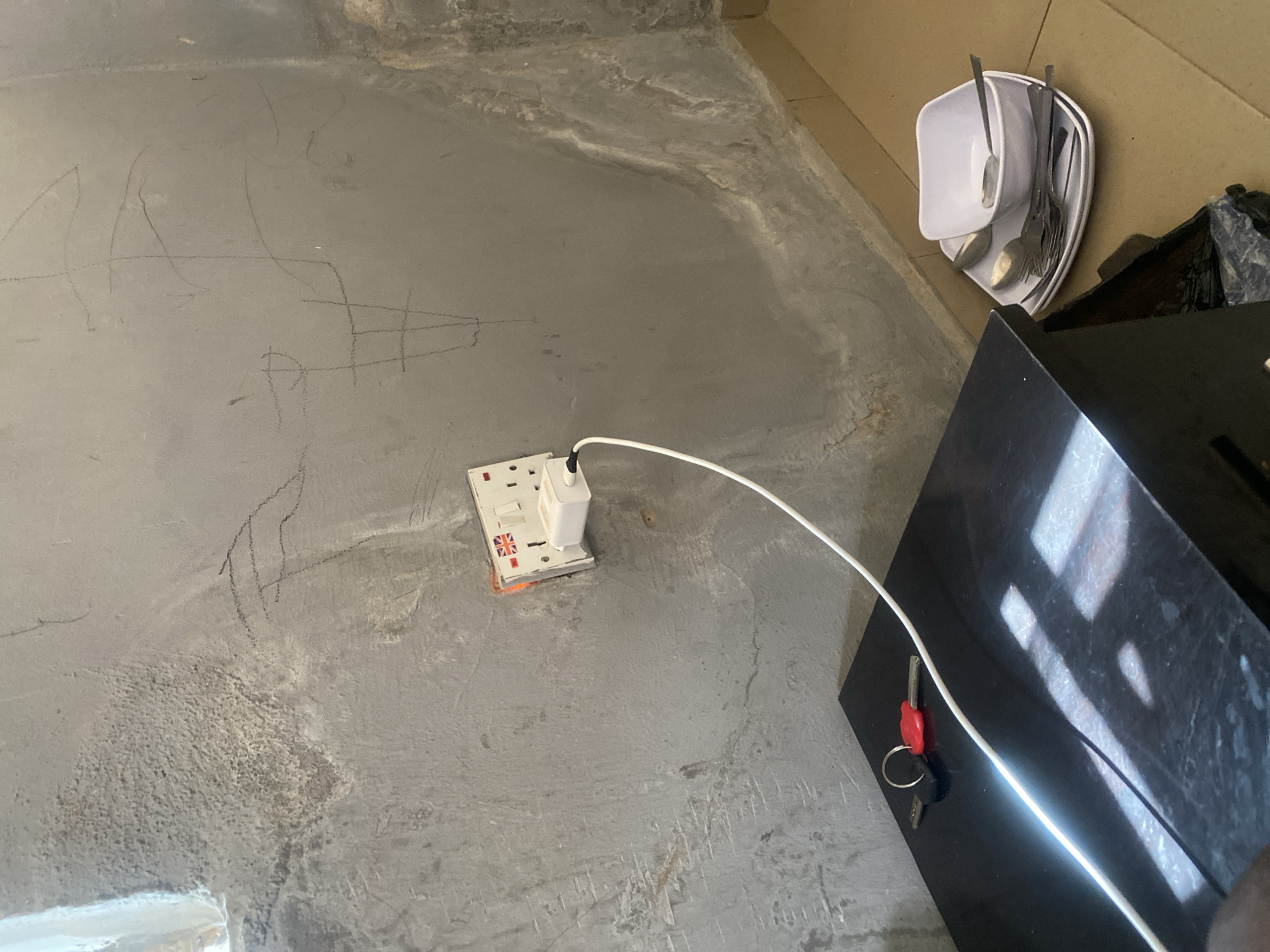This is a medium shot, eye-level, indoor scene taken in Jalingo, Nigeria. The image captures a section of a wall and a bedside table. The wall is a rough, grey, cement-like texture with visible imperfections, water stains, and what appear to be charcoal or pencil drawings. Mounted on the wall is a white electrical outlet with a British flag emblem, and a white plug is inserted. A white cable extends from the plug and runs across the scene.
On the black bedside table, two keys are placed, one with a red head and the other silver. A white cable lies draped across the table. To the right of the table, on the floor, is a stack of white plates and cutlery, including spoons and forks, arranged next to a white bowl. The floor appears to be tiled. The lighting in the scene is somewhat dim, with a strong reflection on the bedside table from an unseen light source.