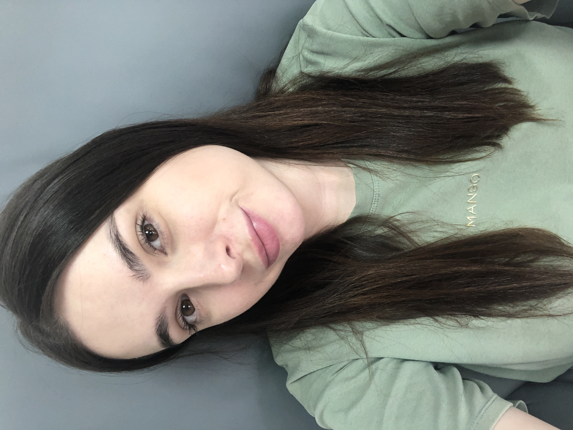 A young woman with long, dark brown hair is reclining on a gray surface, possibly a bed or couch. She is wearing a light green shirt with the word "MANGO" embroidered in gold on the chest. Her face is slightly tilted towards the camera, and she has brown eyes and pink lips. The background is a plain, light gray color. The image was probably not taken in the city of Chaikovsky, Russia, because that information cannot be determined based on the image alone.