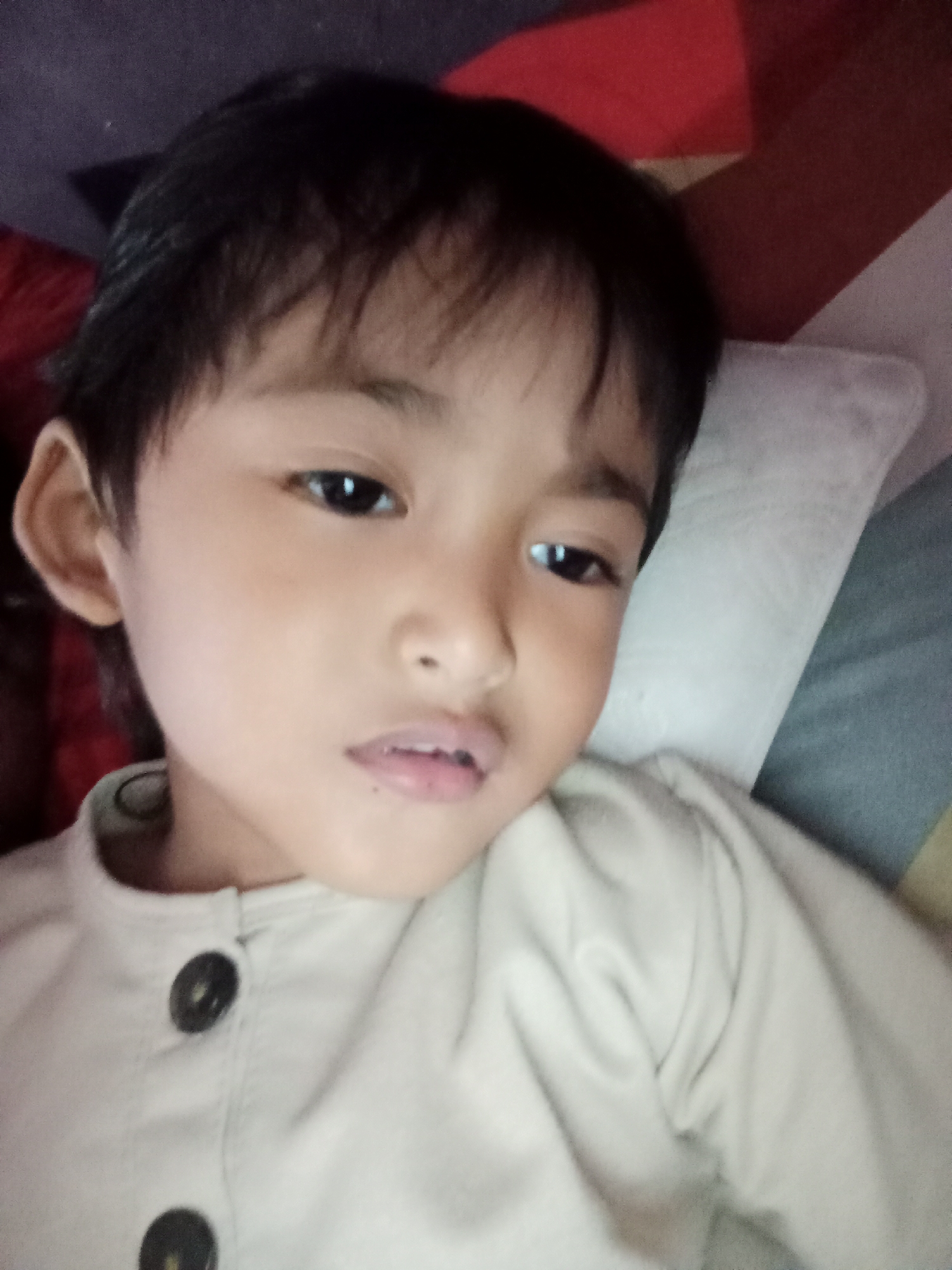 A close-up selfie shows a young East Asian child lying down, resting on a pillow. The child is looking directly at the camera with a neutral expression. Their dark hair is short and slightly tousled. They are wearing a light-colored button-up shirt with two dark buttons visible on the front. The background is blurred but appears to be a bed with patterned sheets or blankets. The image was taken indoors, likely in a residential setting. This image has been geotagged to Bloro, Indonesia.