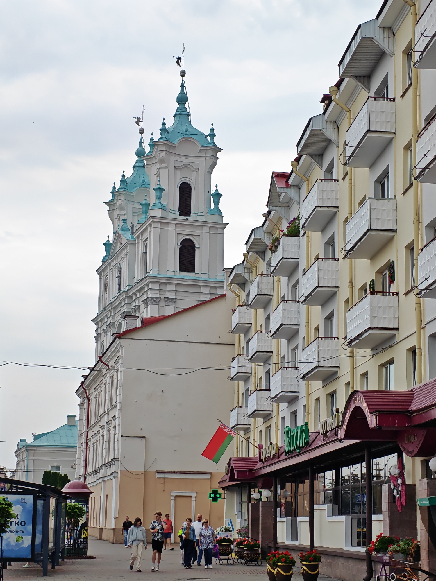 The image depicts a street scene in the city of Hrodna, Belarus, featuring a large, ornate white building with turquoise accents on the left, likely a church or a historical landmark, and a modern apartment building with numerous balconies on the right. The apartment building is painted in a pale yellow color and has a red tiled roof. People are walking on the sidewalk in front of the buildings. A bus stop shelter is visible on the far left. A Belarusian flag is displayed on a pole attached to the apartment building. The sky is overcast.
