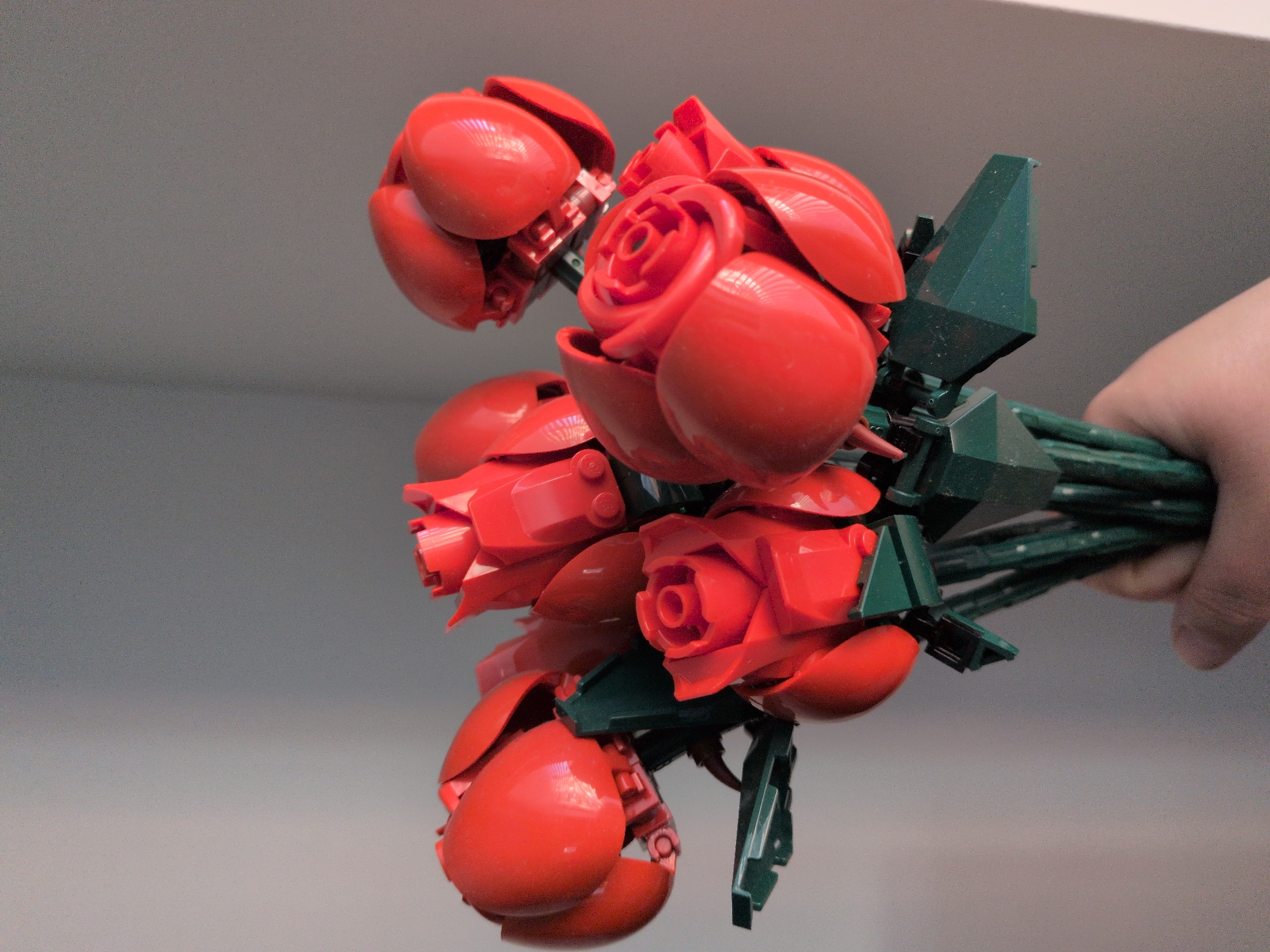 A close-up image depicts a human hand holding a bouquet composed of approximately seven red LEGO roses with dark green stems and leaf elements. The hand, visible at the bottom of the frame with a light skin tone, grasps the bundled dark green LEGO stems and angular leaf structures. The red rose heads consist of multiple smooth, curved red LEGO elements that simulate petals in various stages of bloom, from tight buds to more open forms, revealing some internal red LEGO components. The surface of the red LEGO bricks exhibits reflections, indicating a smooth, glossy texture. The background is a plain, light gray or off-white wall or surface, appearing uniformly lit with some subtle grain. The lighting creates soft highlights on the plastic surfaces. The scene is located in the city of Unknown, Unknown.