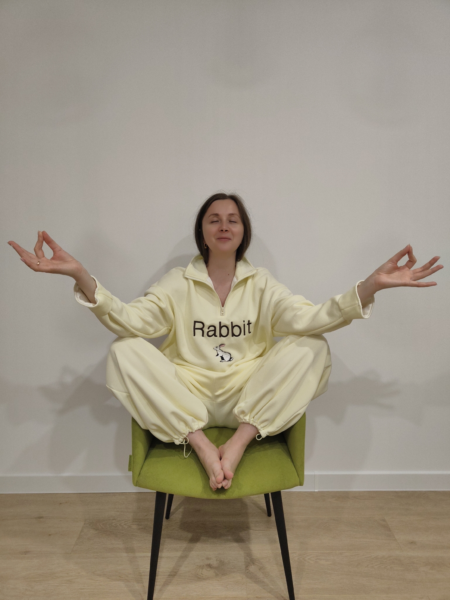 A young woman is depicted sitting in a meditative pose on a vibrant green armchair, against a minimalist background of a plain white wall and a light wooden floor. She is wearing a light yellow (or cream-colored) tracksuit, which features the word "Rabbit" in black lettering on the chest, accompanied by a small black and white rabbit illustration. Her eyes are closed, and her facial expression conveys a sense of peace and relaxation. Both of her hands are raised to her sides, resting on her knees, with her thumb and index finger touching in a classic mudra gesture.
The tracksuit has a half-zip collar and a comfortable, loose fit, with elasticated or drawstring cuffs on the sleeves and pant legs. The green armchair has sleek black metal legs. The setting appears to be indoors, likely a modern room, with soft, even lighting suggesting daytime. The overall impression is one of tranquility and calm.