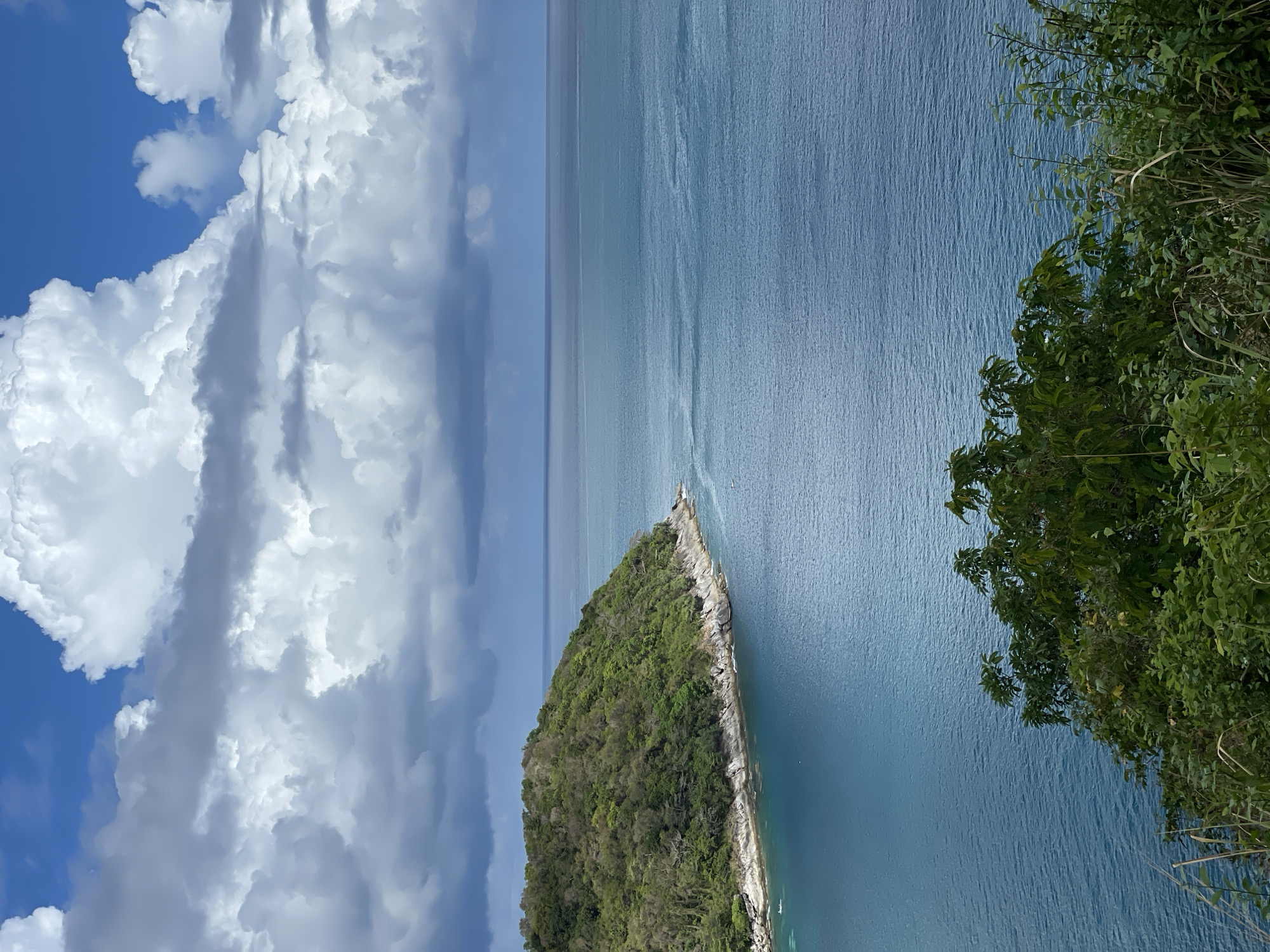 The image depicts a coastal scene, likely taken from an elevated vantage point, possibly near the city of Rawai, Thailand. In the foreground, green foliage cascades from the right side of the frame, suggesting a cliffside or steep slope. A triangular-shaped island, densely covered with trees, extends into a calm body of water. The water transitions in color from a lighter, clear blue near the island to a darker, deeper blue towards the horizon. The horizon line is straight and clear, separating the water from the sky. Above the water, a dramatic cloudscape fills the left side of the frame, featuring large, puffy white clouds against a patch of clear blue sky. The overall atmosphere suggests a sunny day with scattered cloud cover. There are no people or animals visible in the scene.